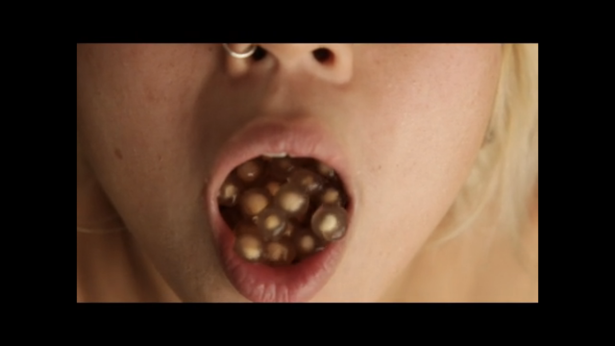 A still from a video depicting the lower half of a person's face, mouth agape filled with translucent tan seeds or tapioca balls.