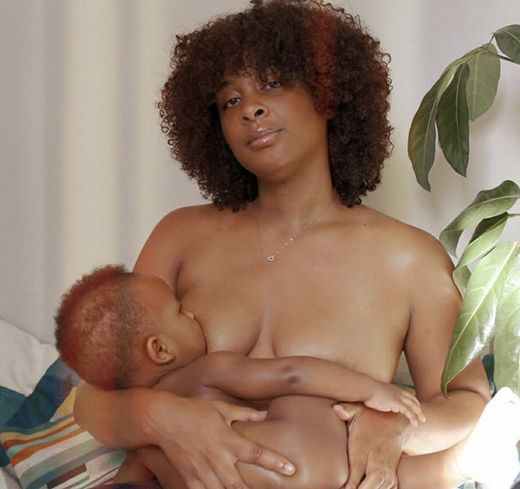 A mother breastfeeding her child, both nude, her curly hair covering a portion of her face as she confidently stares into the camera.
