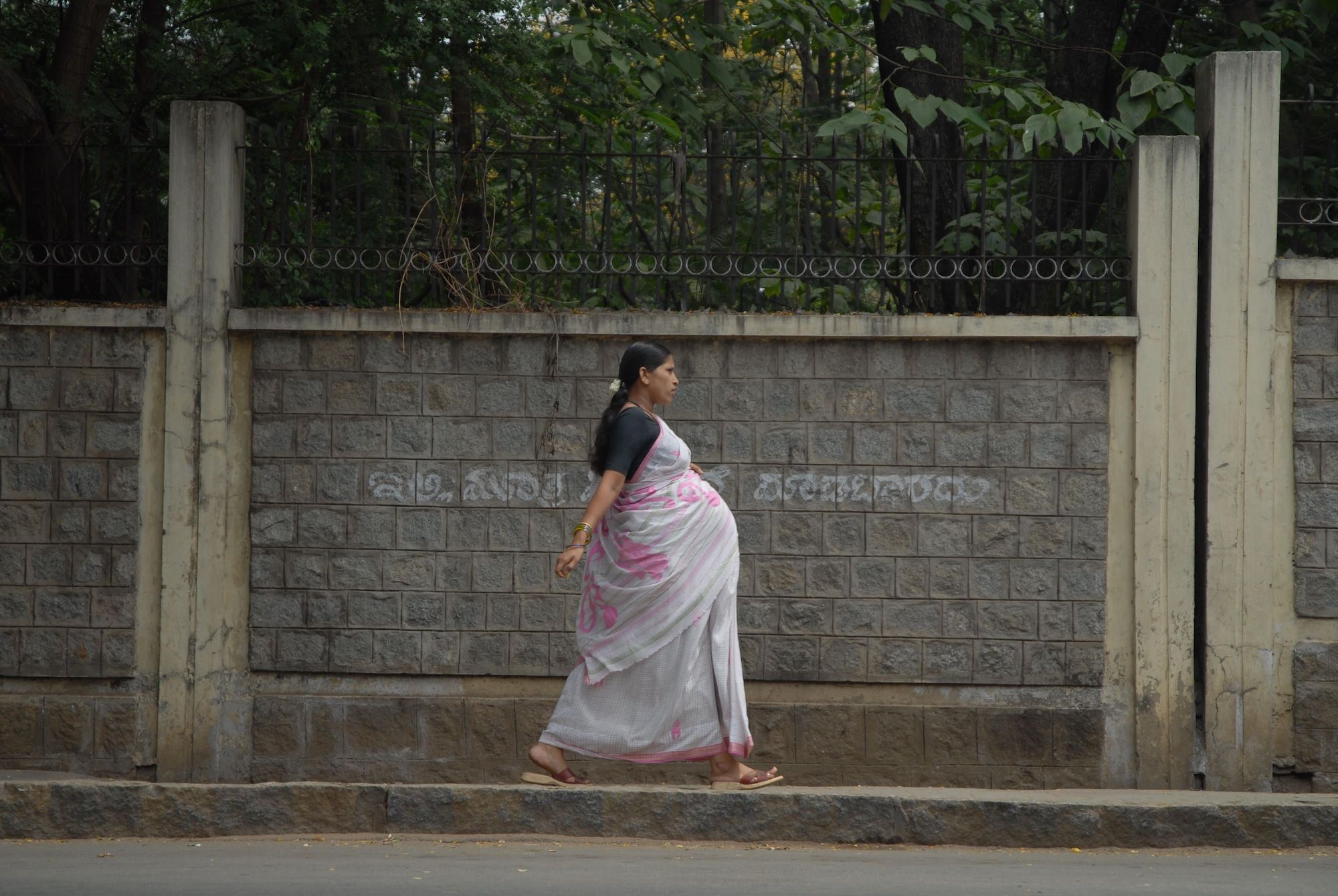 A visibly pregnant woman walking along a sidewalk against a gray concrete backdrop with green tress looming up above.