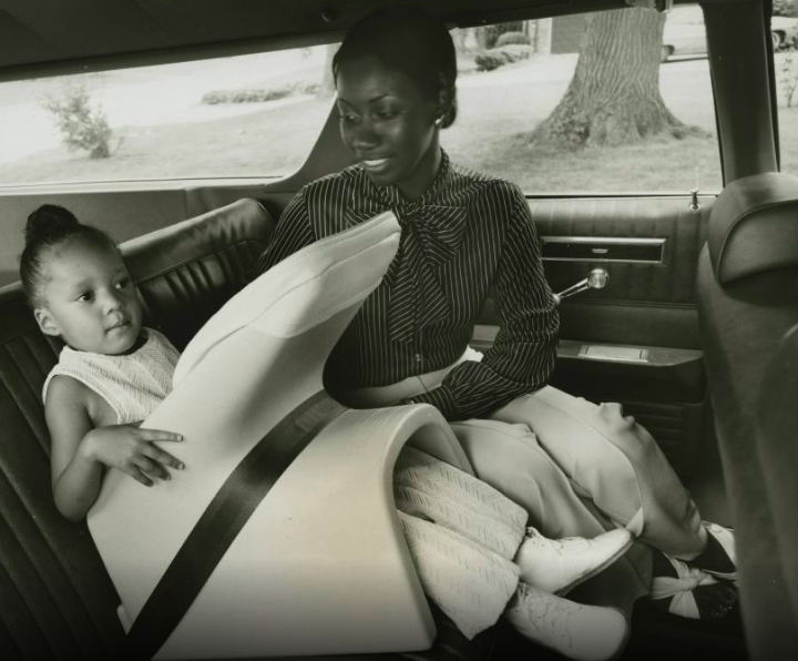 A black and white photograph of a woman and her child in the backseat of a car; the child is sitting in a futuristic looking car seat to protect her.