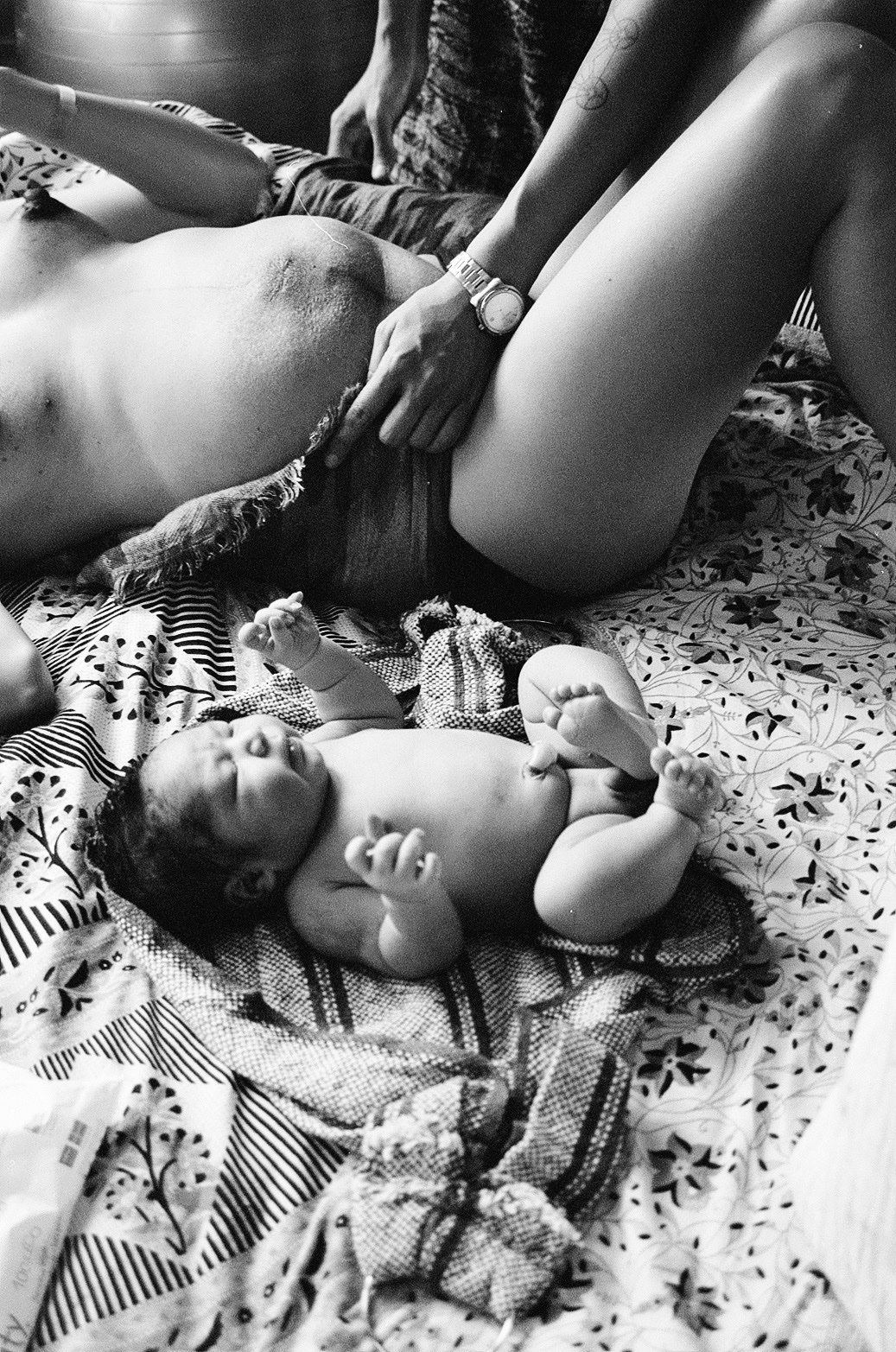 A black and white photo of a baby laying belly up in the foreground and what is speculated to be its mother in the background in the background, being wrapped up by a pair of hands that appear from the top right corner of the image.