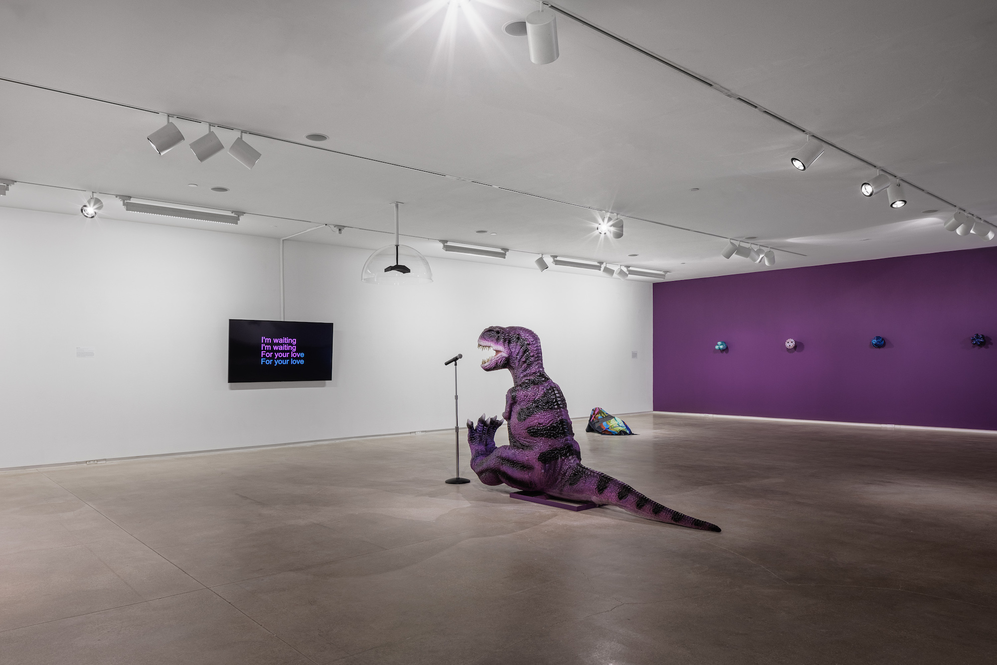 The far wall, a lush purple color of what is for the most part a white cube gallery but in the middle a T.Rex sits in the middle of the room in front of a microphone and tv, reading karaoke lyrics.