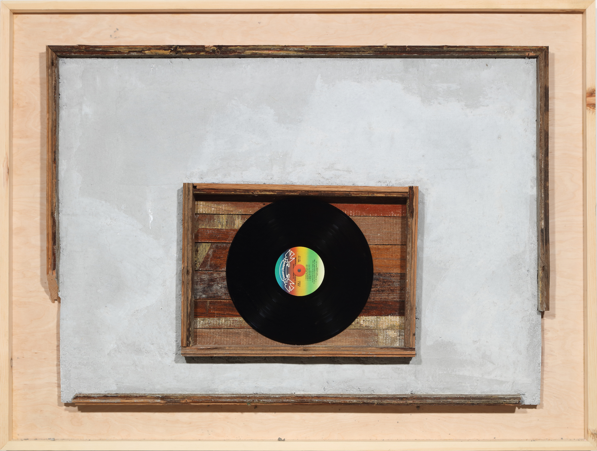 A rectangular panel of wood housing a gray slab of concrete hung horizontally with a smaller wooden panel and a vinyl record embedded in the middle of the slab.