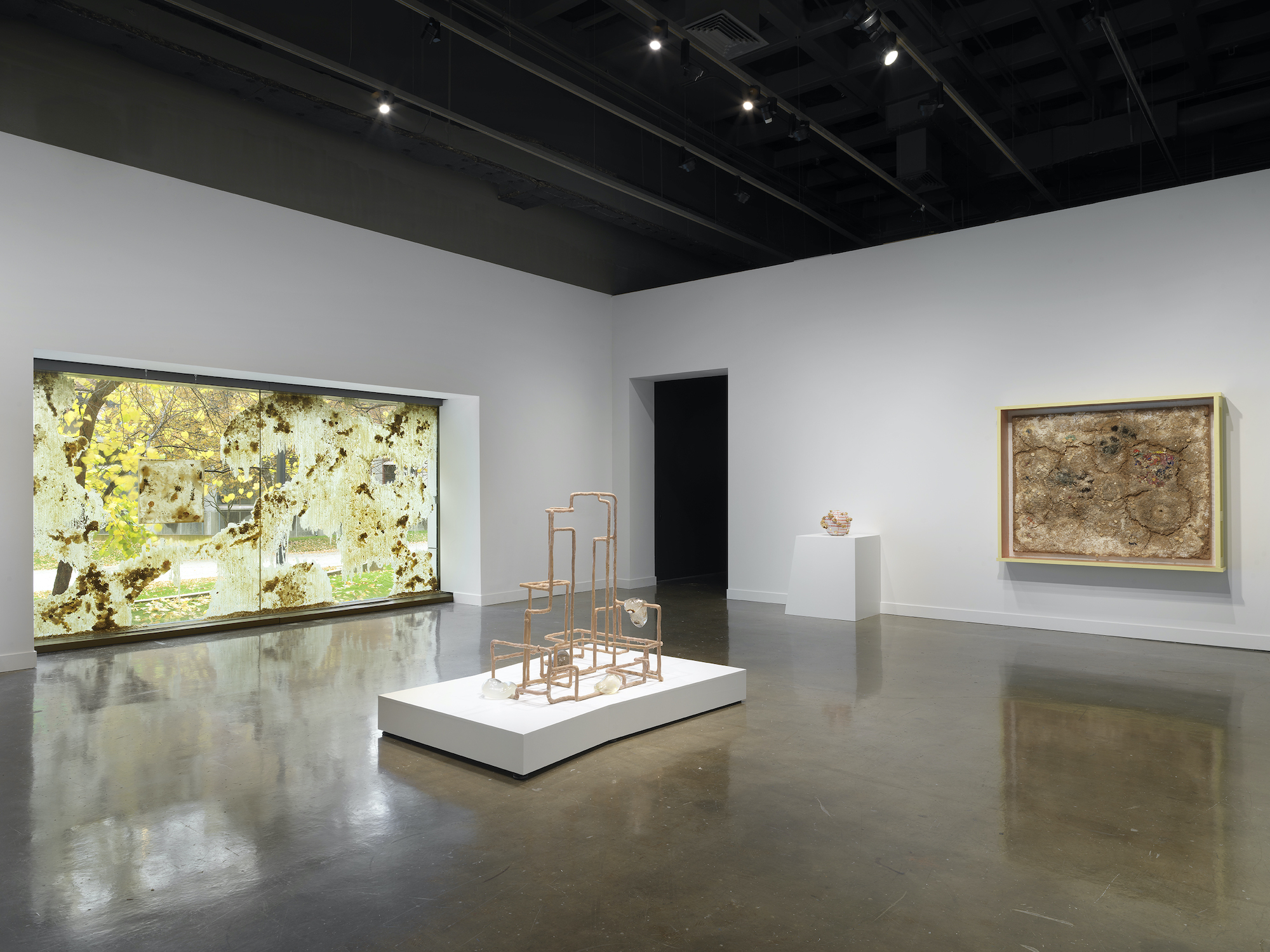 A window installation of drips and smears of beeswax and flora, behind a geometric sculptural work in the center on a white platform. A smaller spherical sculpture sits on a white pedestal and a large rectangular sculpture is hung on the wall.