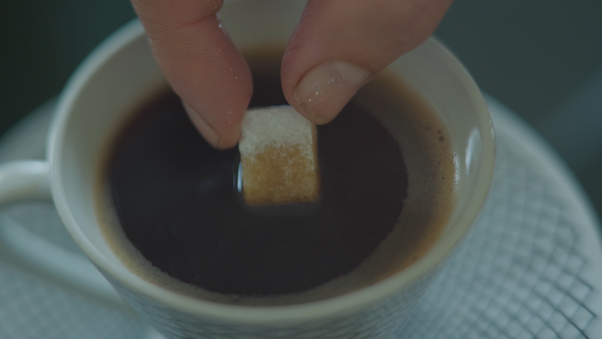 View of hand pinching a sugar cube, dipping it into a coffee mug containing a dark liquid