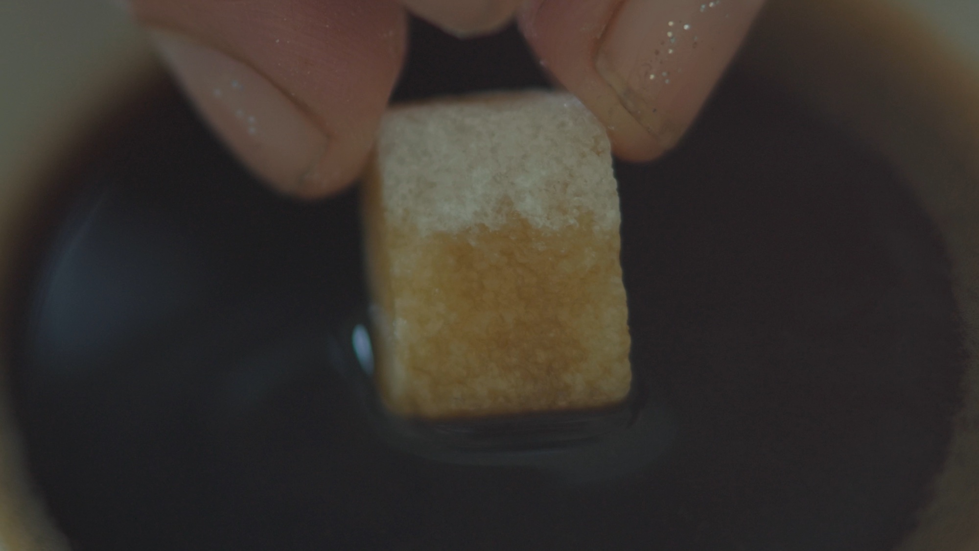 Close up view of a hand pinching a sugar cube, dipping it into a dark liquid