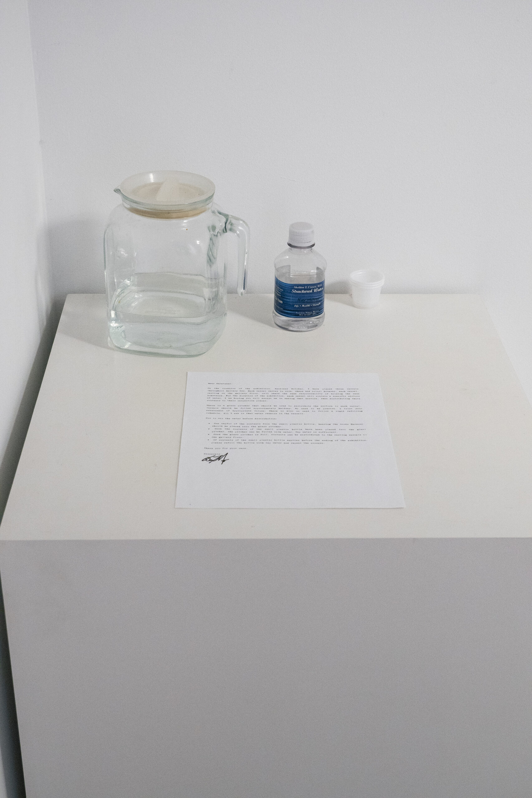 A glass vessel holds water, and sits next to a water bottle, which is half full. In front of these objects, a typed note sits.