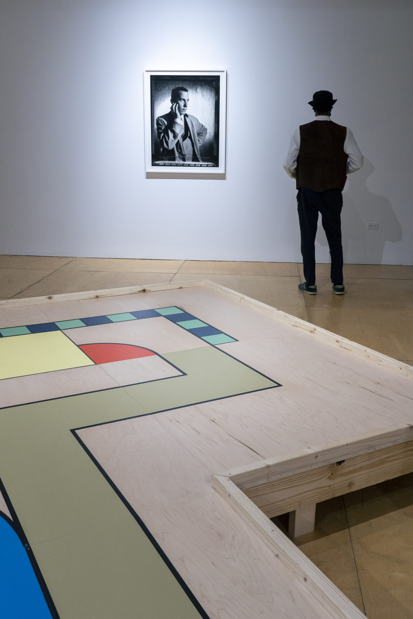 Large wooden platform with colorful geometric shapes sits in a room, adjacent to a portrait of a man in black and white. An onlooker observes the wall.