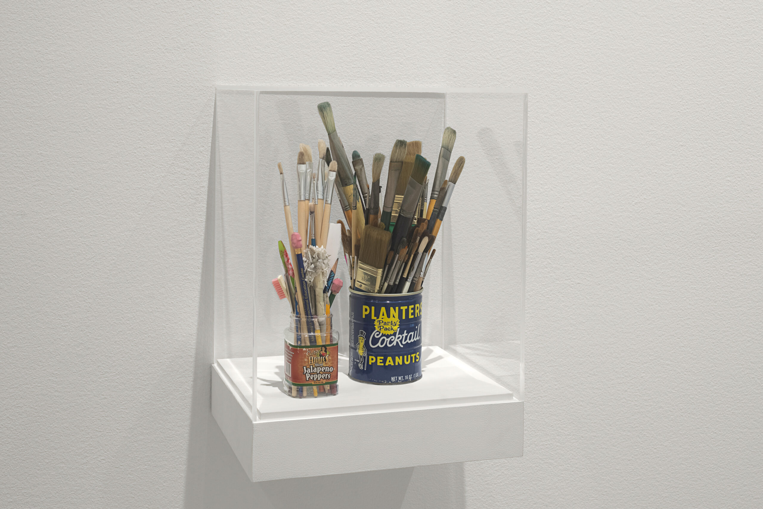 Two cans of paintbrushes housed within a glass box atop a stolid white shelf.