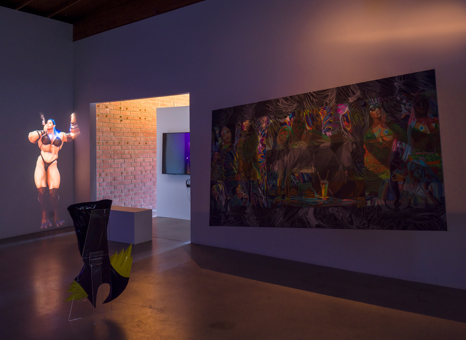 A dimly lit room shows a large digitally rendered femme body builder projected upon the wall with a claw-like sculpture in front of it and a crooked rectangle of various colors on the wall perpendicular to it.