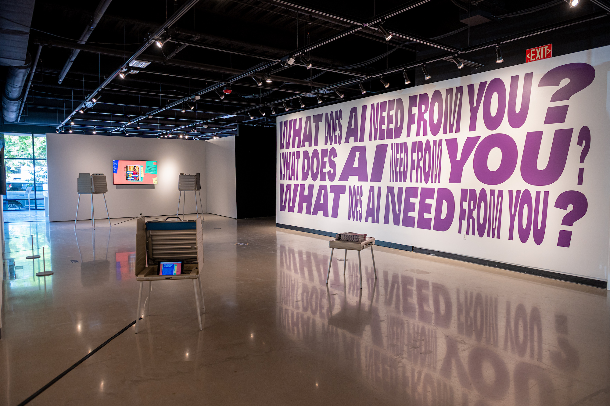 A gallery with a shielded desk with monitor resembling a voting station, text on the wall in the background reads, 
