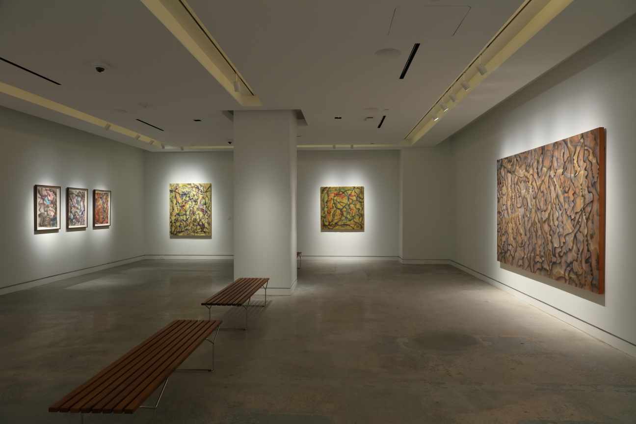 A gallery space with two wooden benches in the middle, paintings adorning white walls.