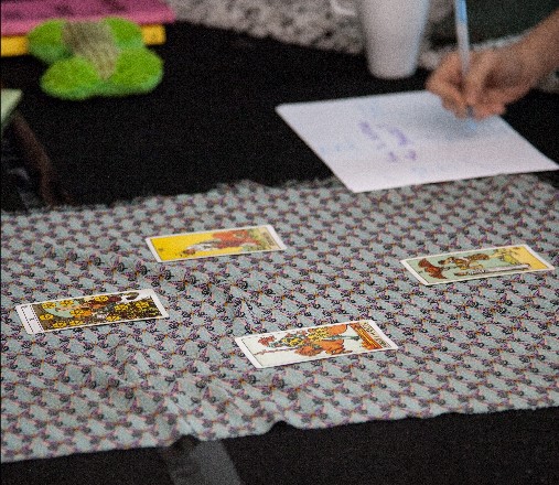 A tarot card reading being done with four cards laid out on top of a table cloth, a hand writes down something on paper in the background