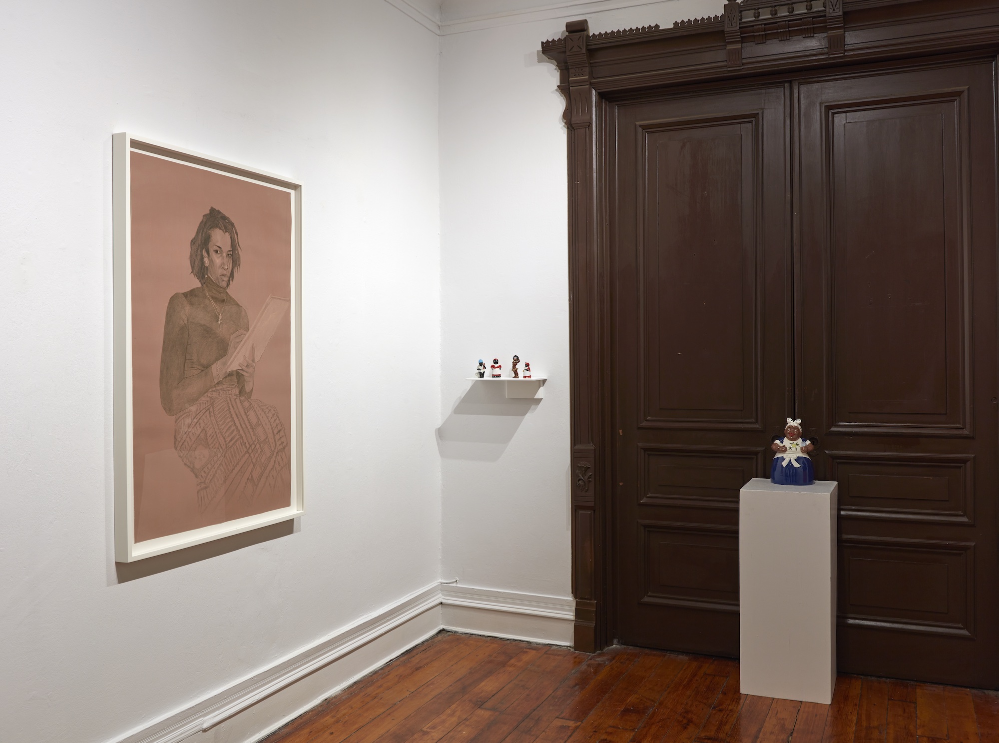 Image of a women sitting down with a book in her hand and sculptures sitting to the right of her in front of a brown door
