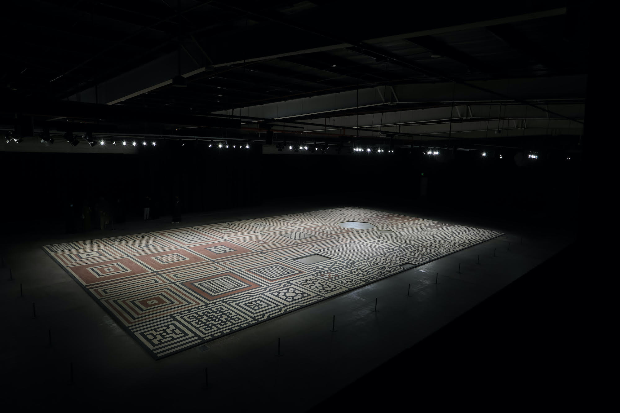 Image of a large empty room with a spotlight shining down onto a mat with many different geometric designs and squares in black, white, and terracotta.