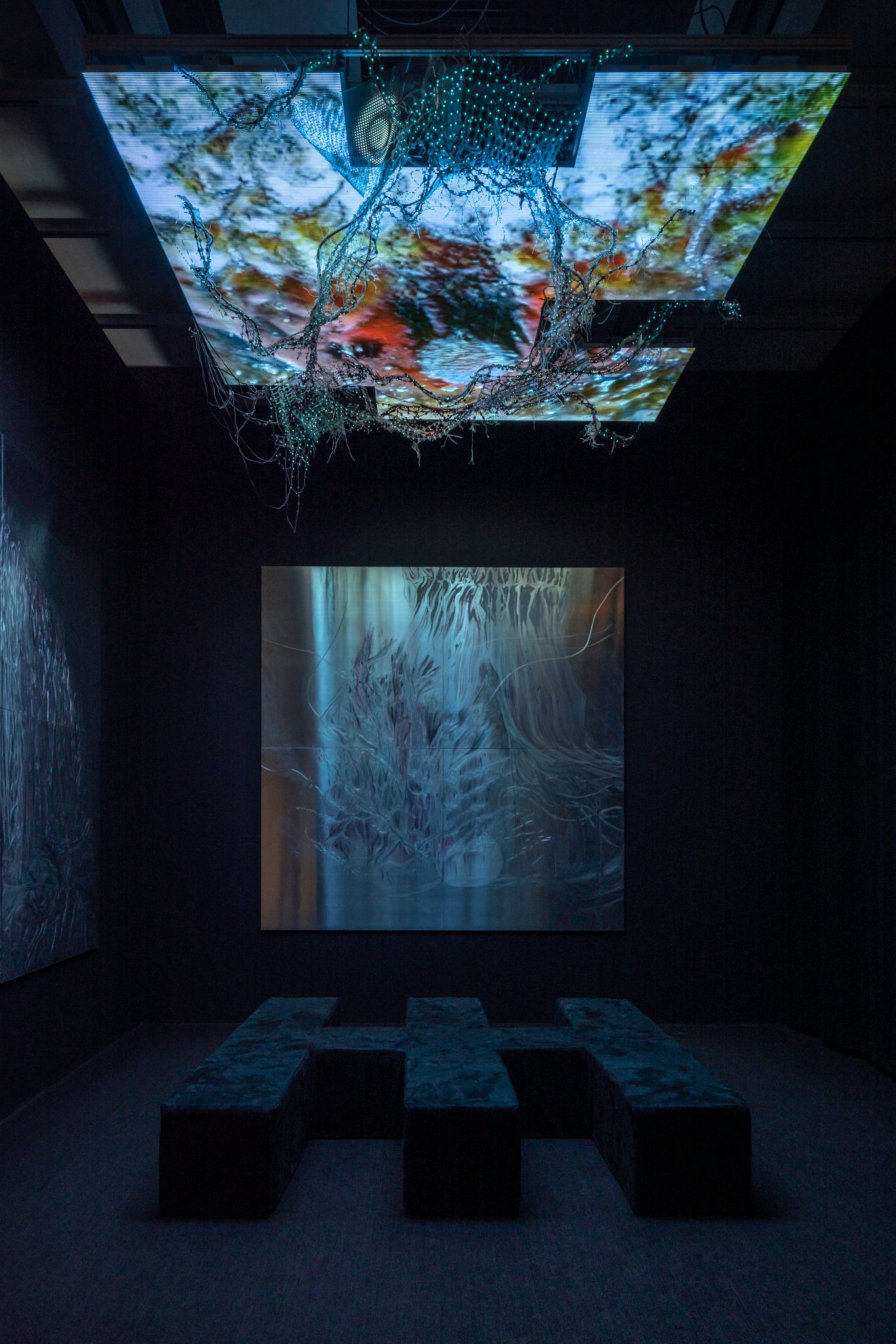 Image of a dark room with a projected screen of swirling blues at the back of the room and a lattice-style bench in the middle. Above the bench is a blue and green speckled screen with protruding blue netting.