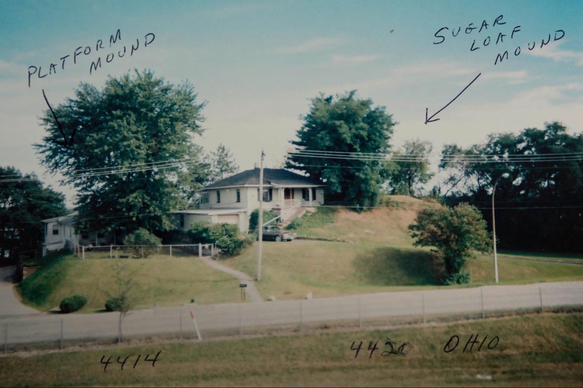 A scrapbook photo of a house between trees on top of raised earth, with handwritten labels stating 