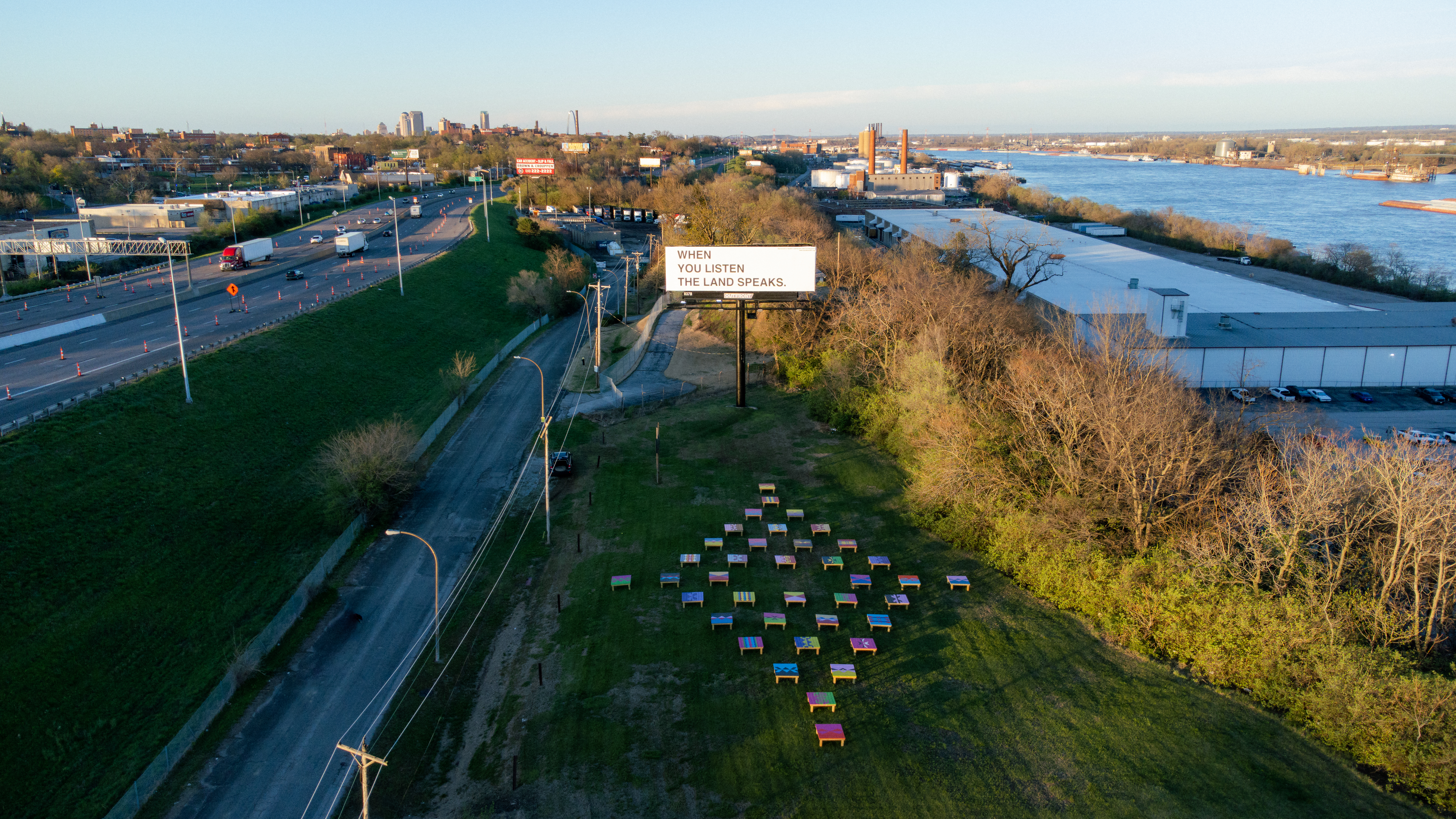 An aerial photo of a green space between a highway and a river, with colorful wooden structures laid out in the shape of a diamond. Above is a white billboard with the text, 