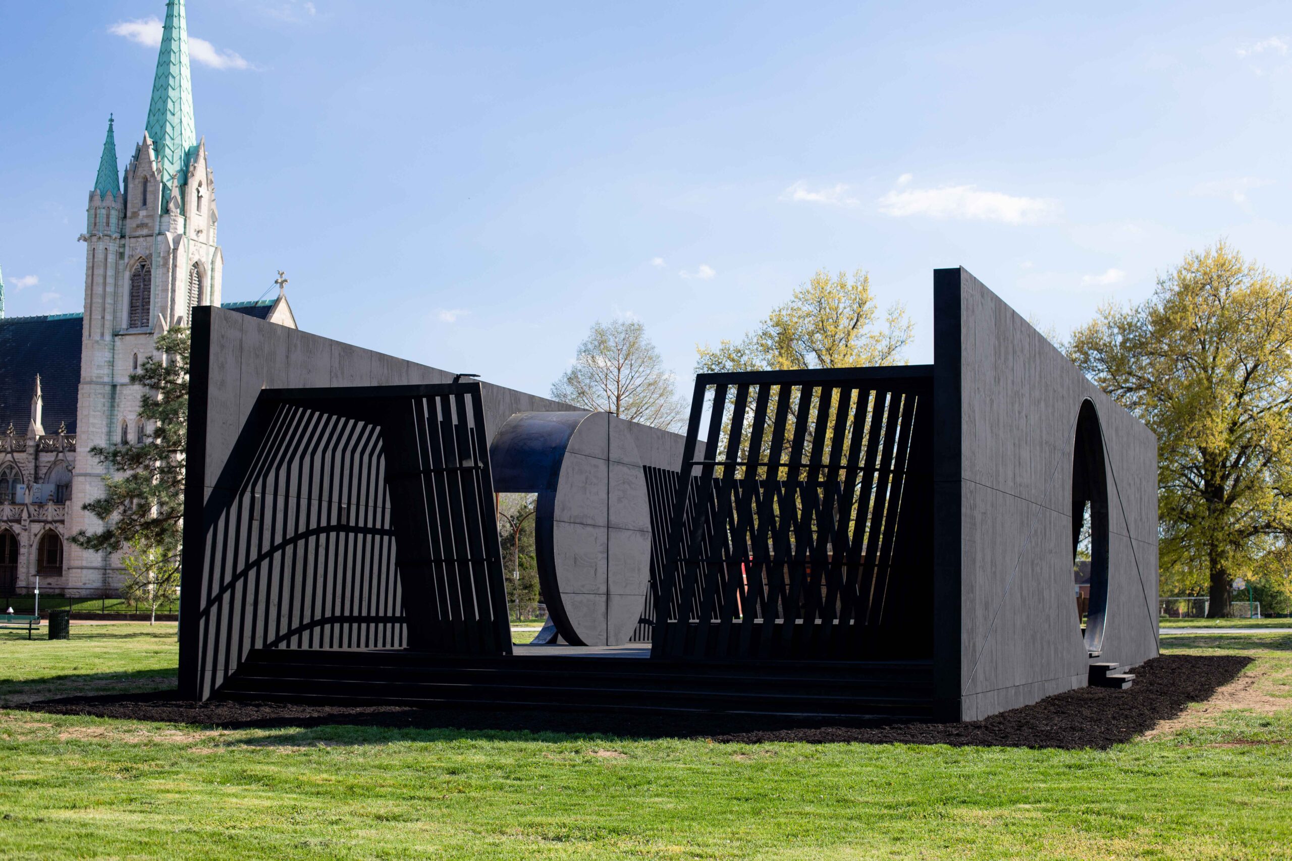 A photo of a black geometric wooden structure located on a public green space, with a church in the background.