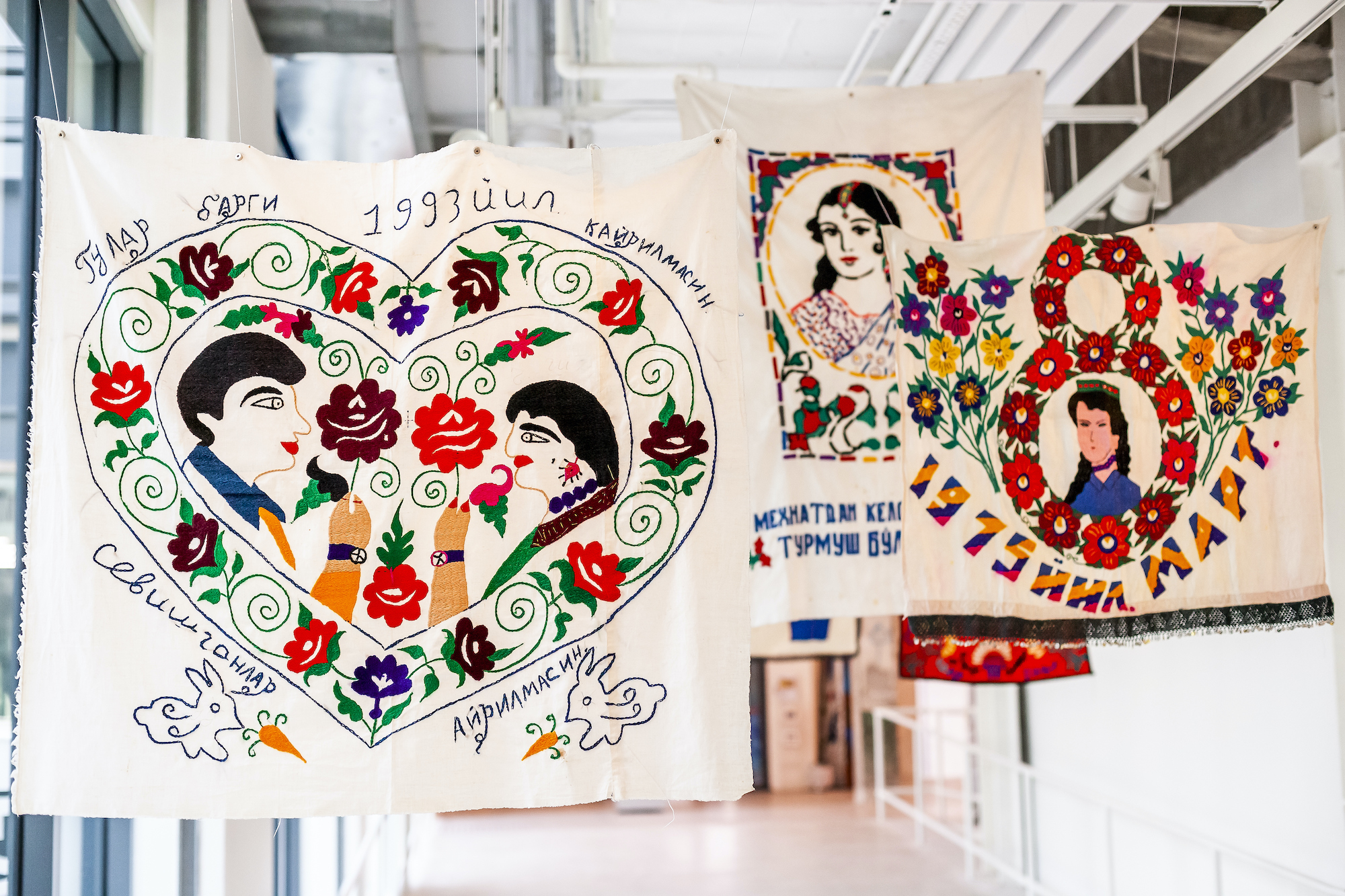 Image of several pieces of fabric hanging from the ceiling, each embroidered with colorful images of people and flowers.