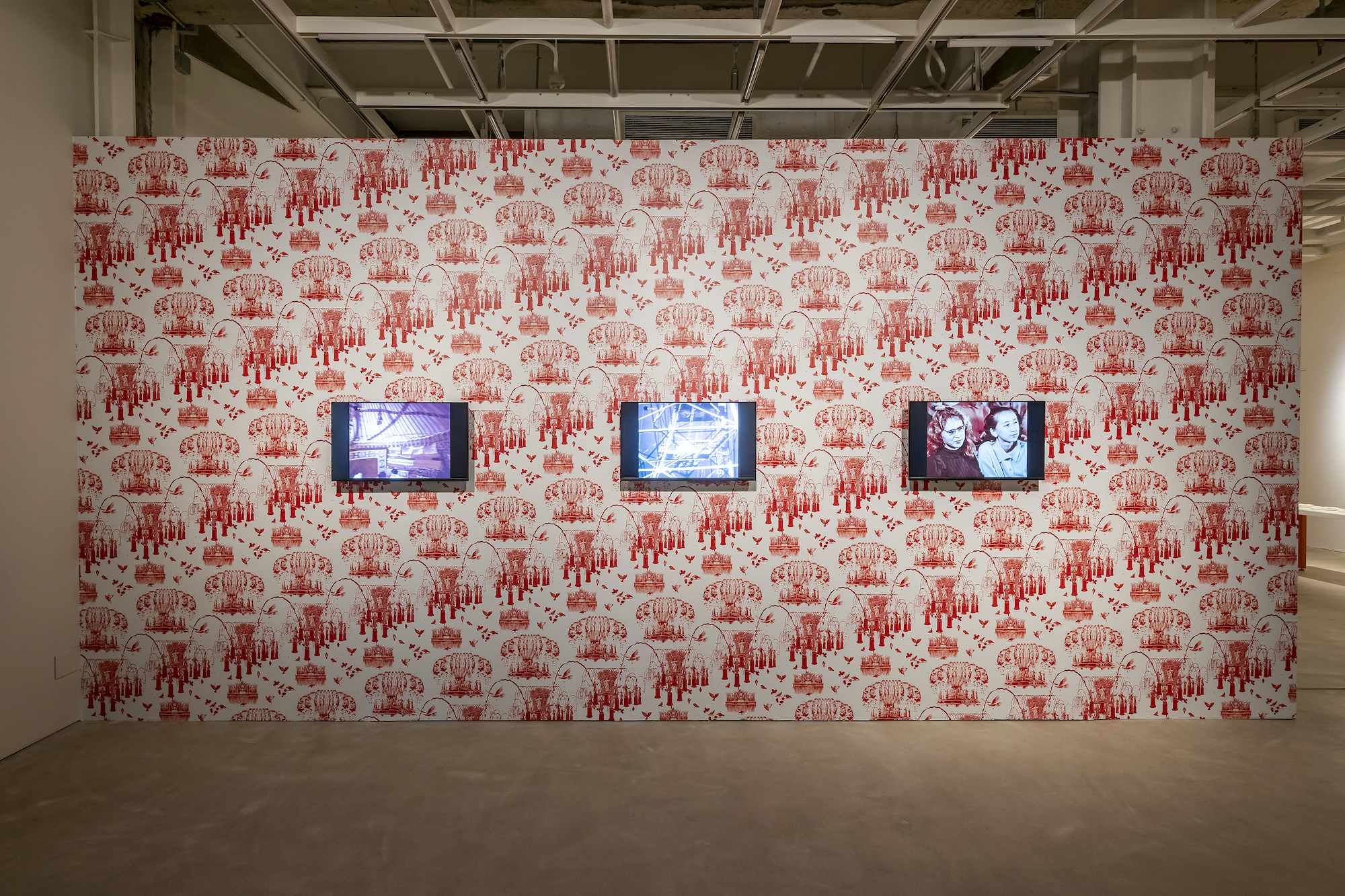 Image of a red and white patterned wall with three small screens organized in a line. The first screen shows a purple-toned image, the second blue, the third red with two figures looking to the right.