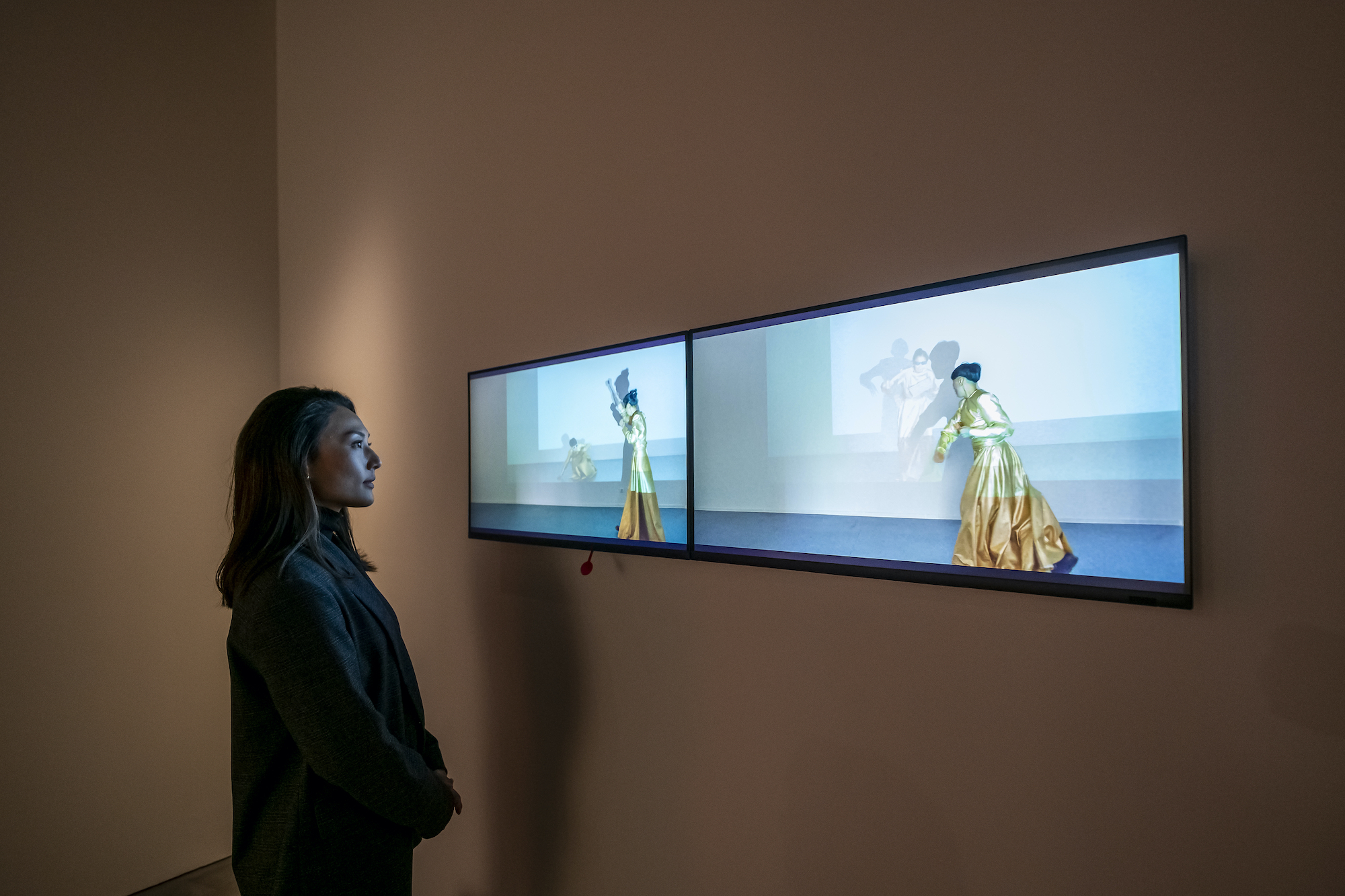 Image of a woman who's face is lit by the two screens she is watching in a dark room. The screen shows a blue background and a figure in a long yellow and orange dress.