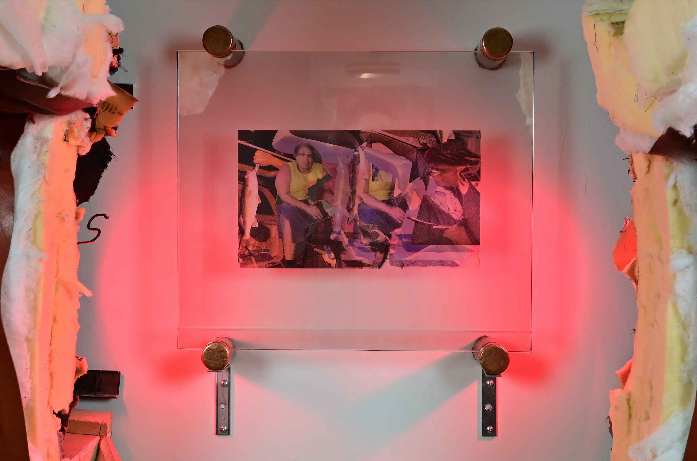 Image of a photograph collage behind a glass pane affixed to a white wall. Shining onto the glass is a hot pink light.