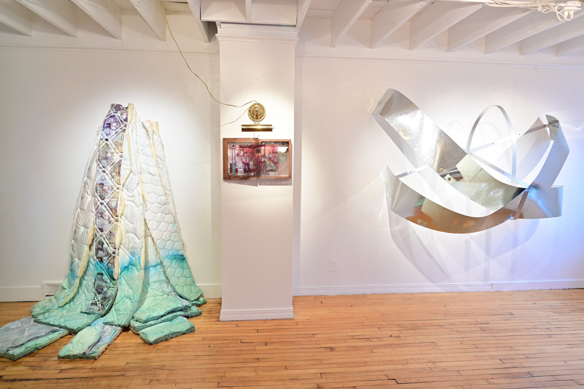 Image of a mattress cut into strips, partially dyed blue, and hung on the wall. Next to it are several silver metal sheets arranged in a swooping way. Between the two is a framed image with a light fixture shining onto it.