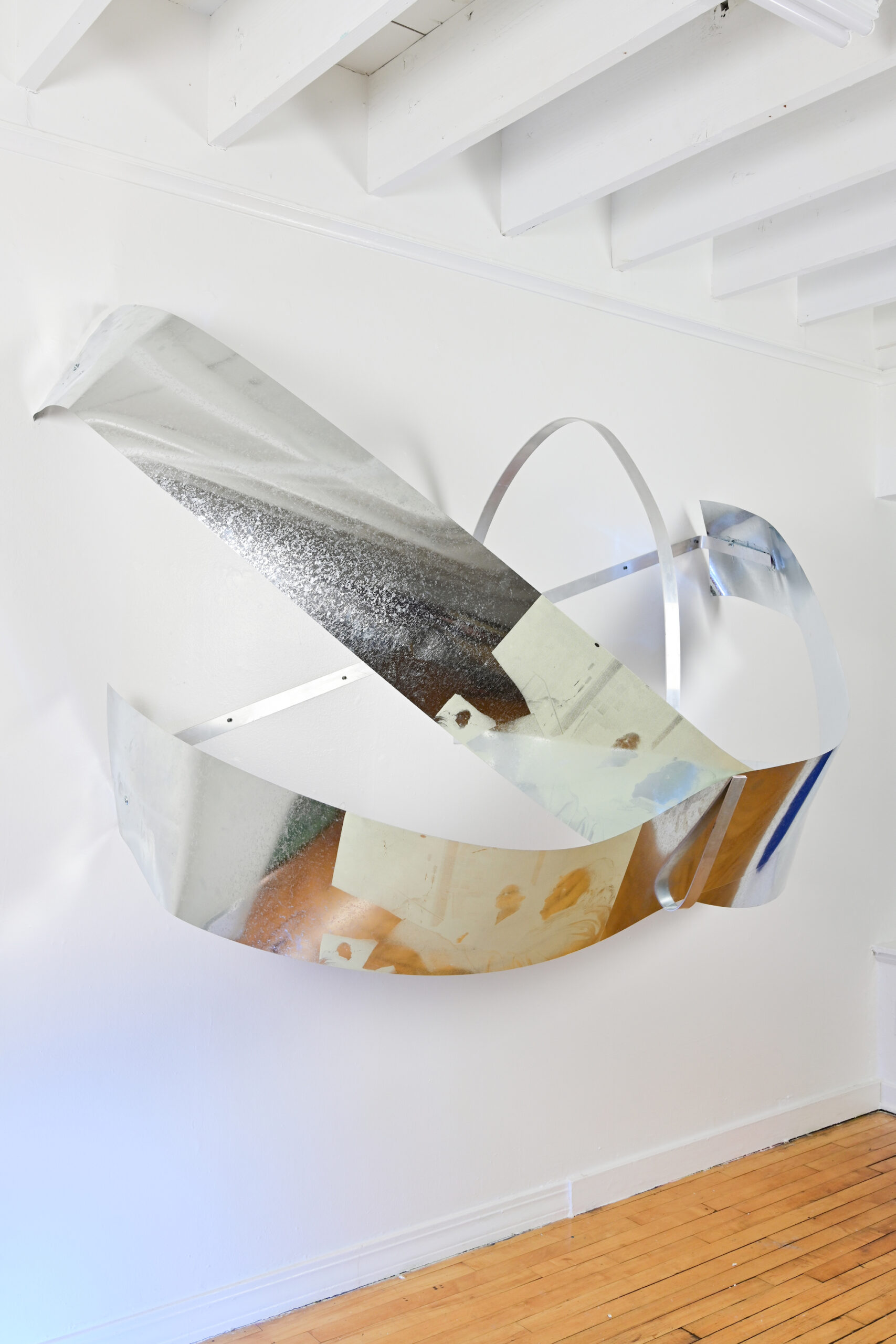 Image of a silver sculpture affixed to a white wall. The sculpture is made of silver metal sheeting cut into thick and thin strips and arranged in a curved manner.