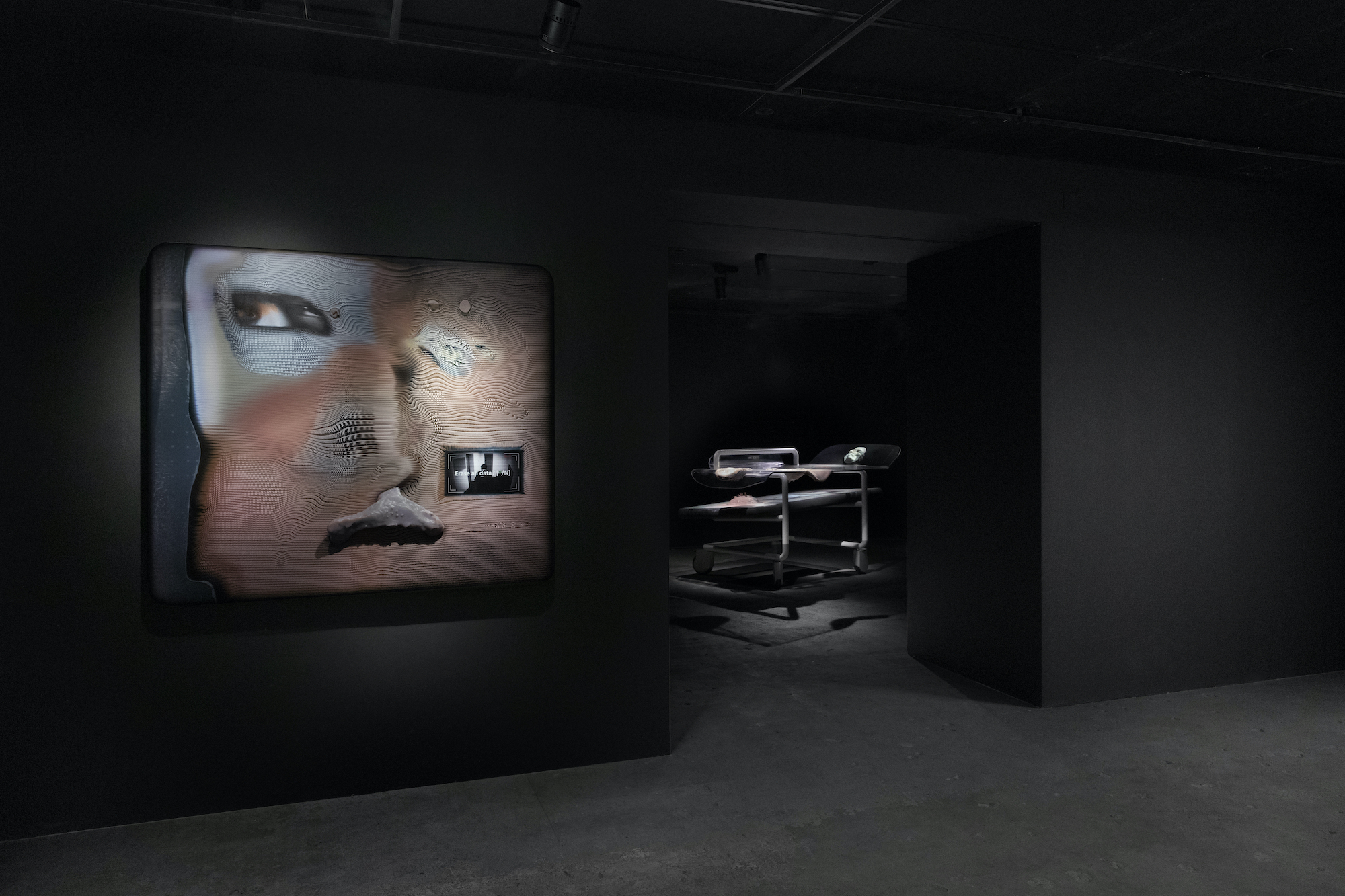 Image of a dark room with black walls and floor featuring a blue and red face-like artwork. Through a doorway is a spotlighted metal cart.