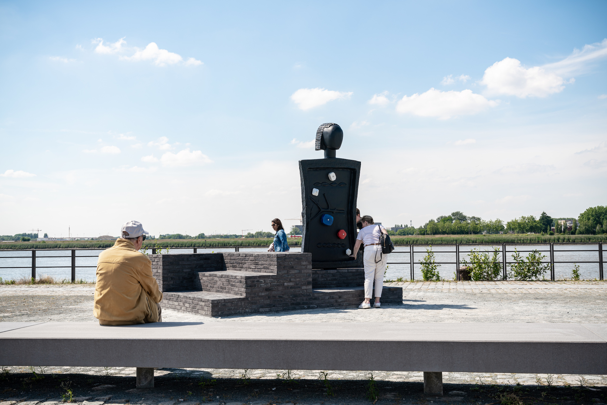 A person on a bench in front of a large figurative / monumnetal sculpture resides next to a river front.