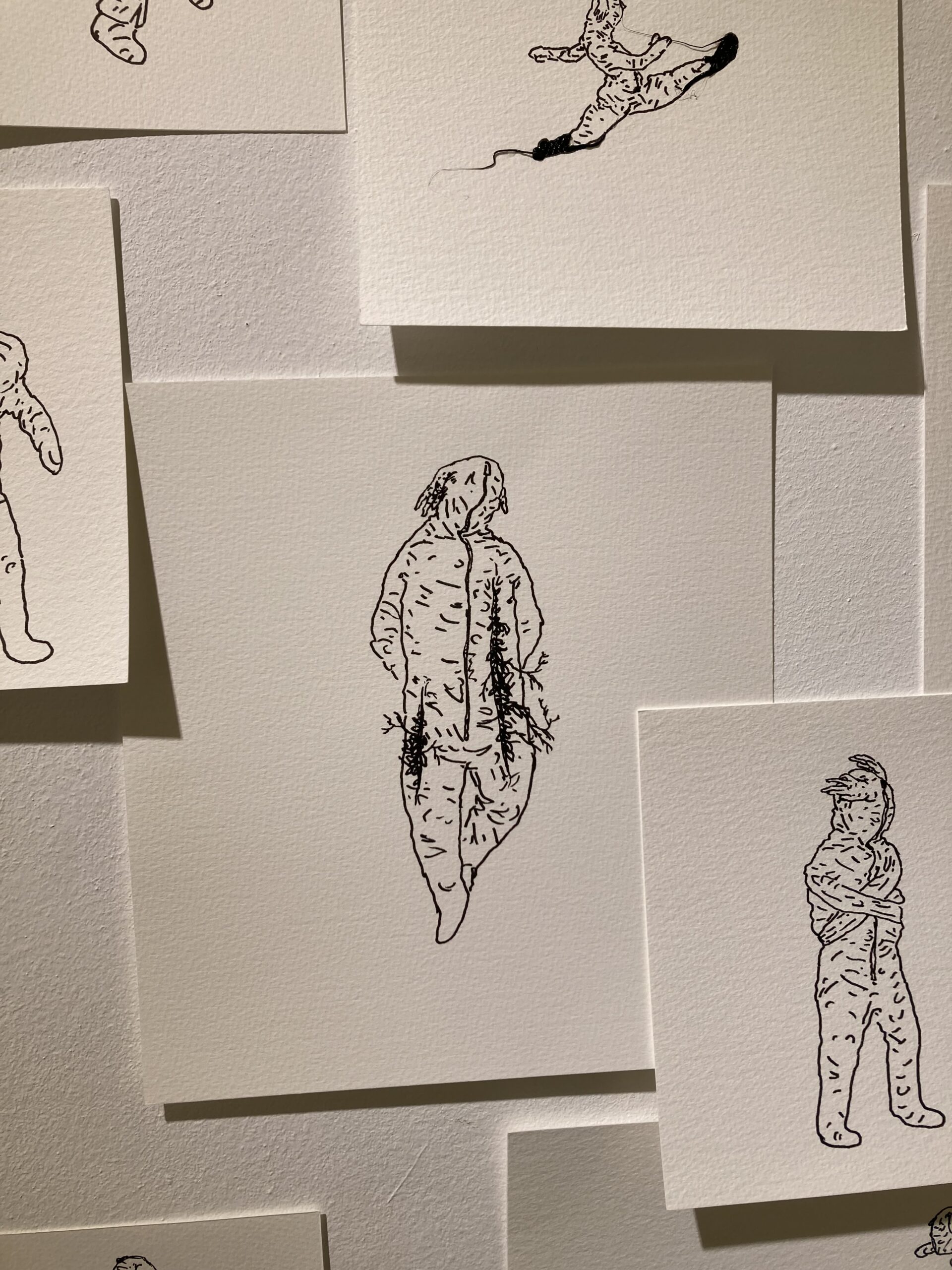 A black and white drawing of a figure on a square piece of card stock surrounded by other drawings posted on a white wall.