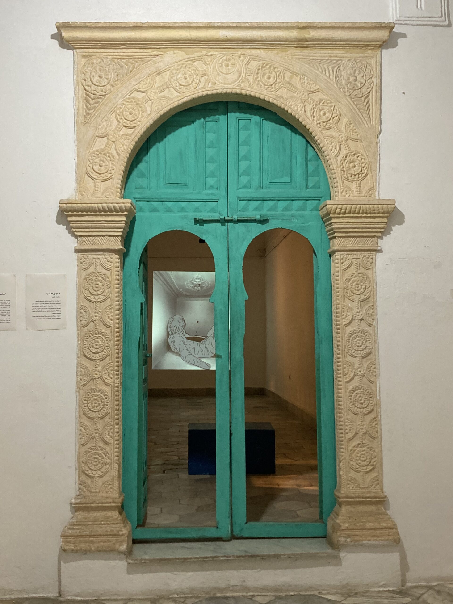 Image of a off-white carved archway in front of a teal blue double door, behind which is a room with a projected screen on the wall.