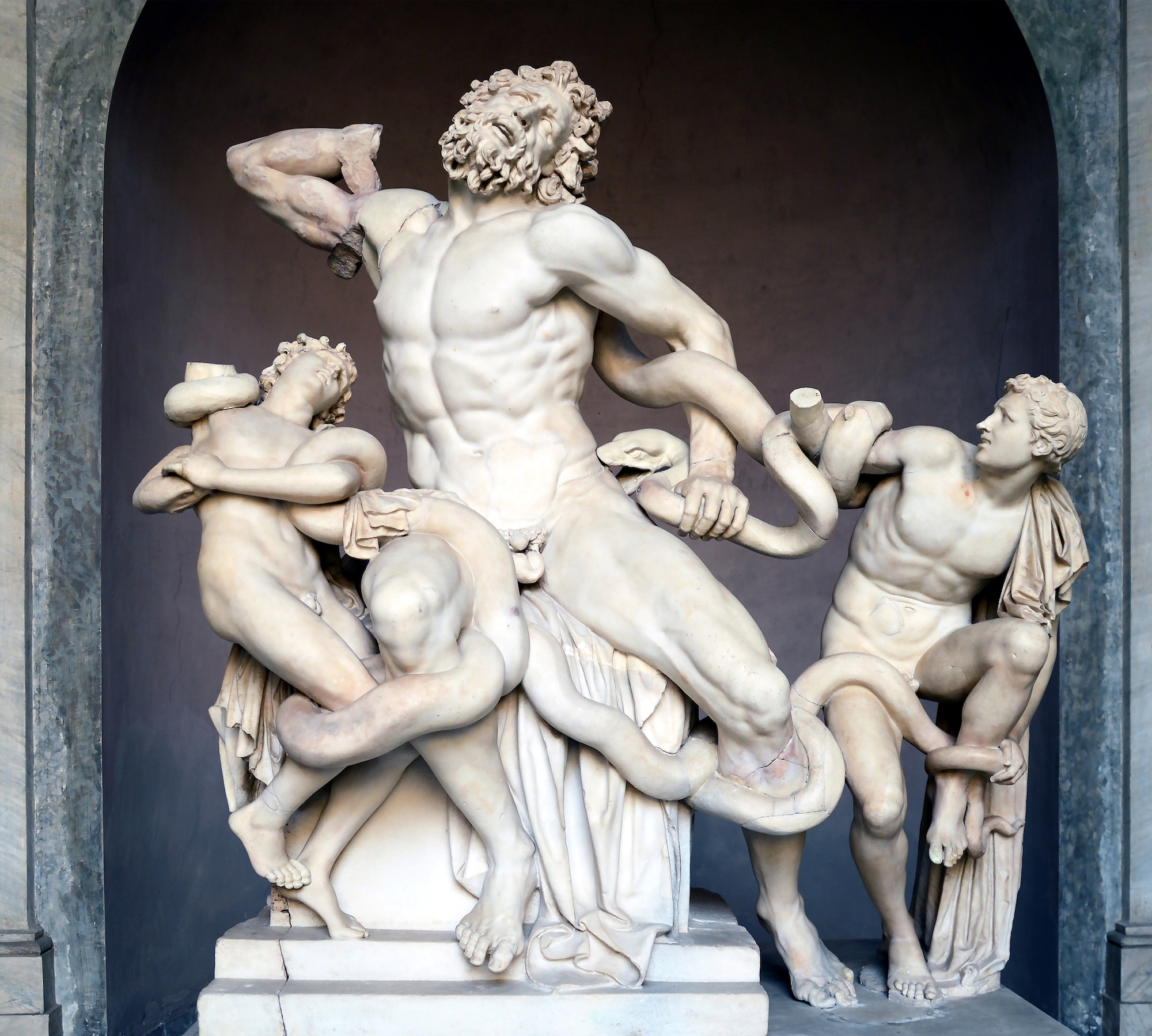 A white marble statue of the mythical Laocoon group displaying a muscular bearded man with two smaller figures poised on either side and snakes writhing between all of them.
