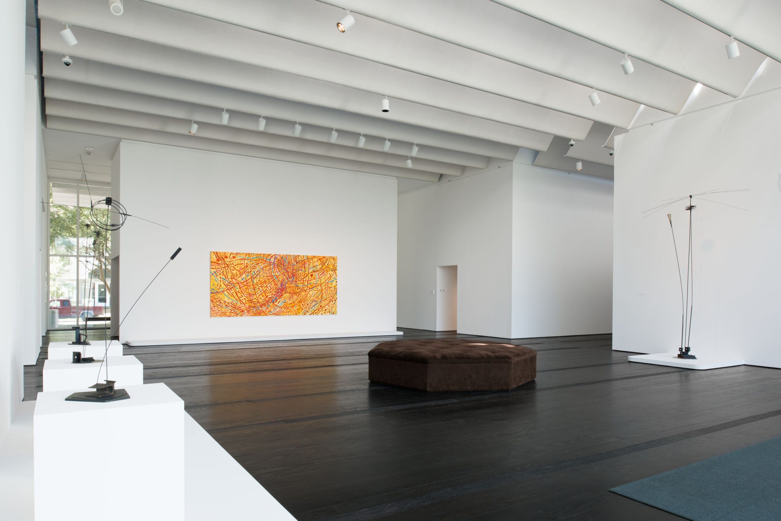 Image of a gallery space with white walls and black flooring featuring a yellow, red, and blue rectangular painting and several tall, dark wire sculptures.