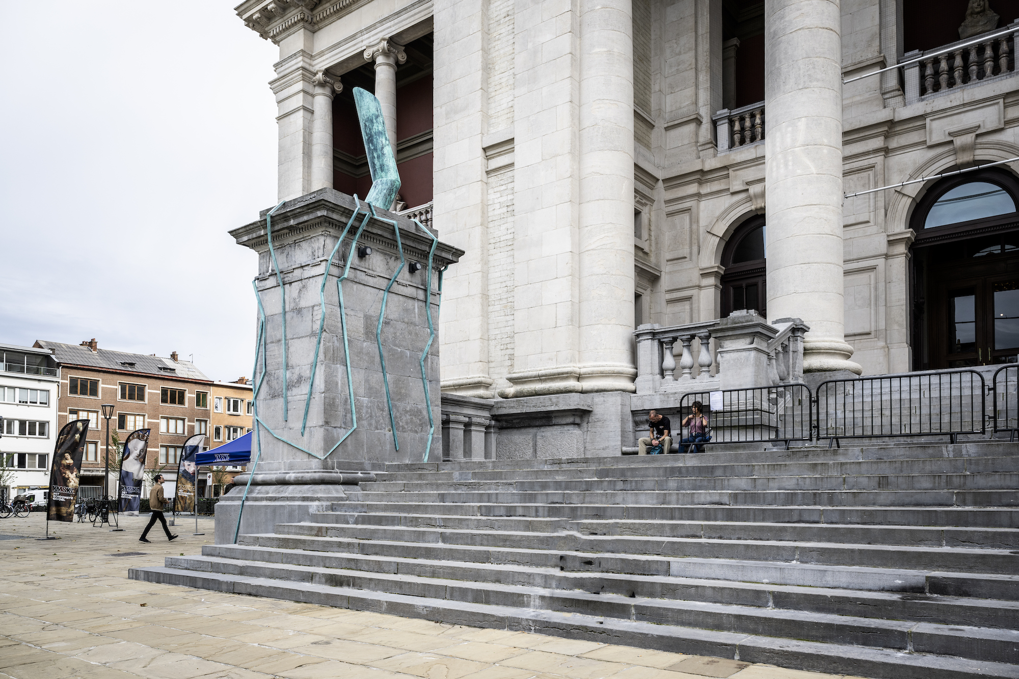 There are steps next to a monumental sculpture leading up to a large museum with giant columns