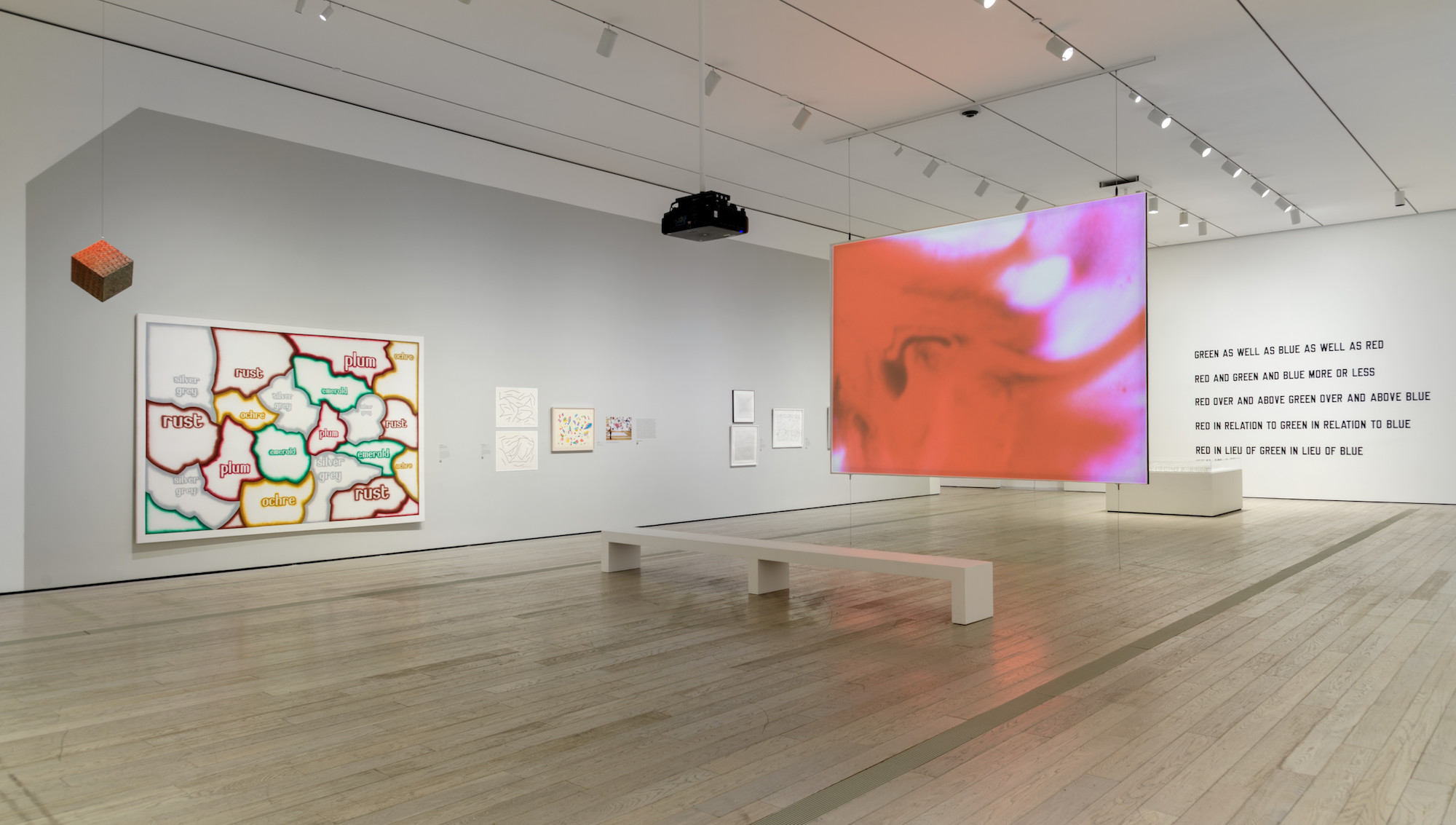 A view of the gallery with smooth tan wooden floors, a large colorful map hangs on a wall as well as large wall text on the far end of the gallery.