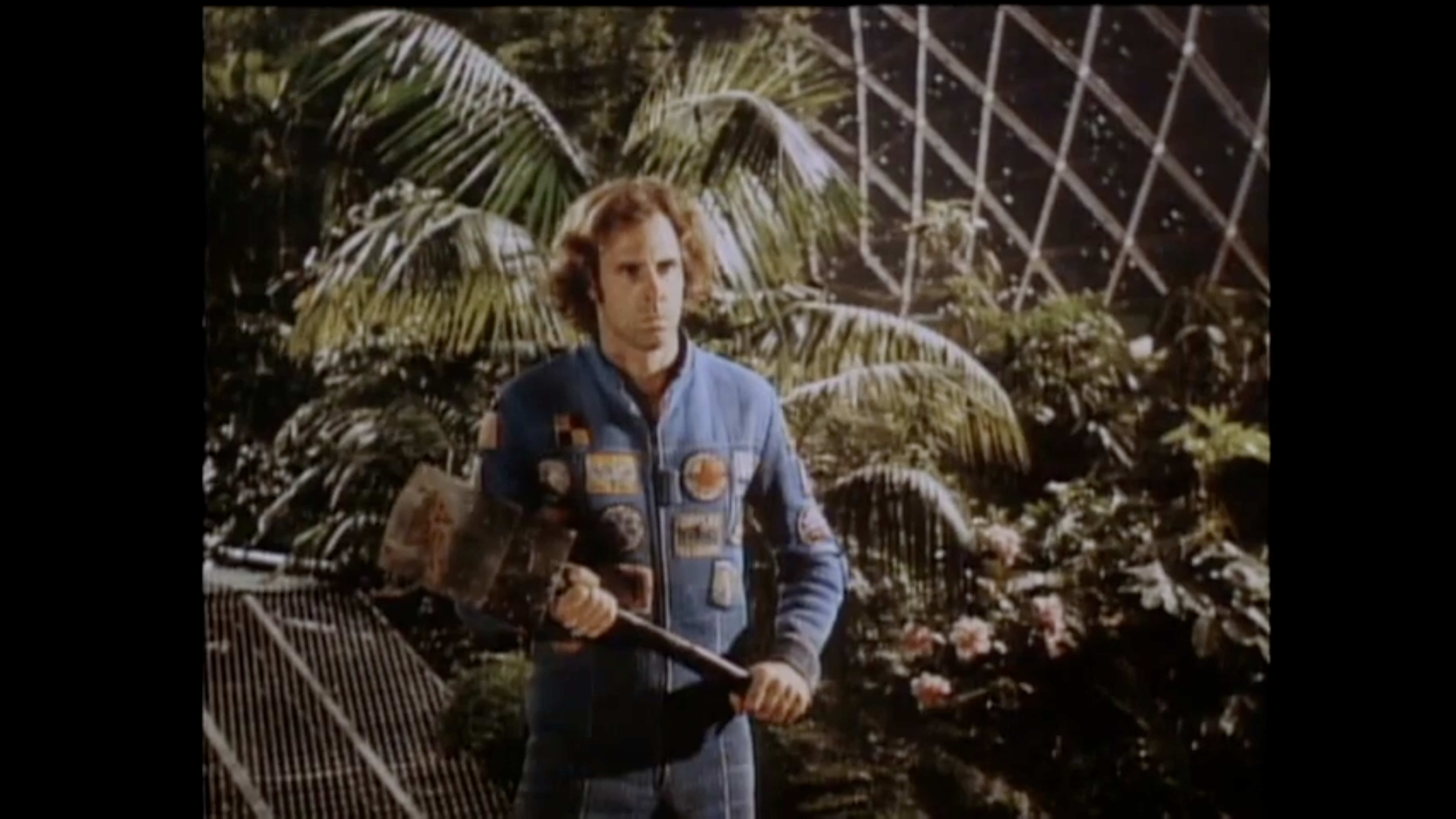 A screenshot of a video of a man walking menacingly with an axe in hand, in front of a background of lush garden within a domed structure.