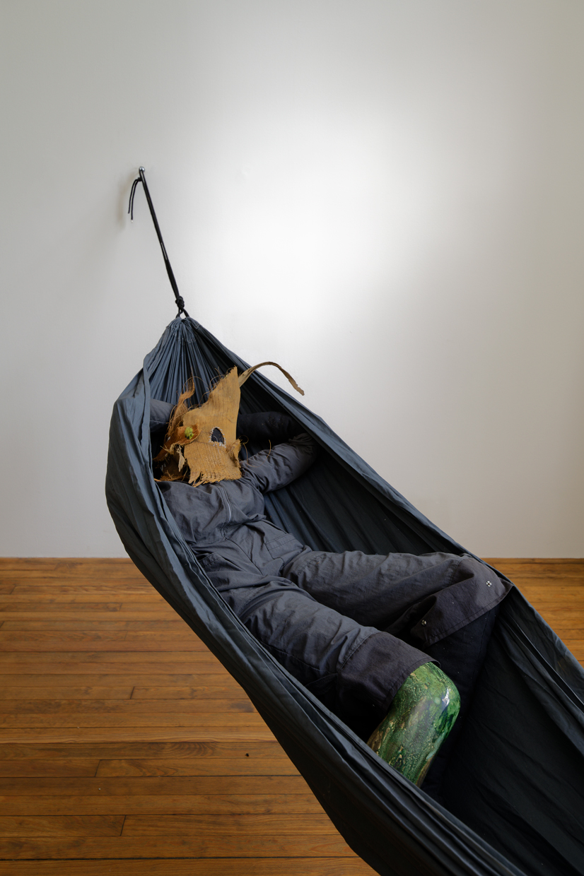 In a white room hangs a hammock with a stuffed figure made of overalls, palm bark, fiber mask, and a ceramic leg laying inside it