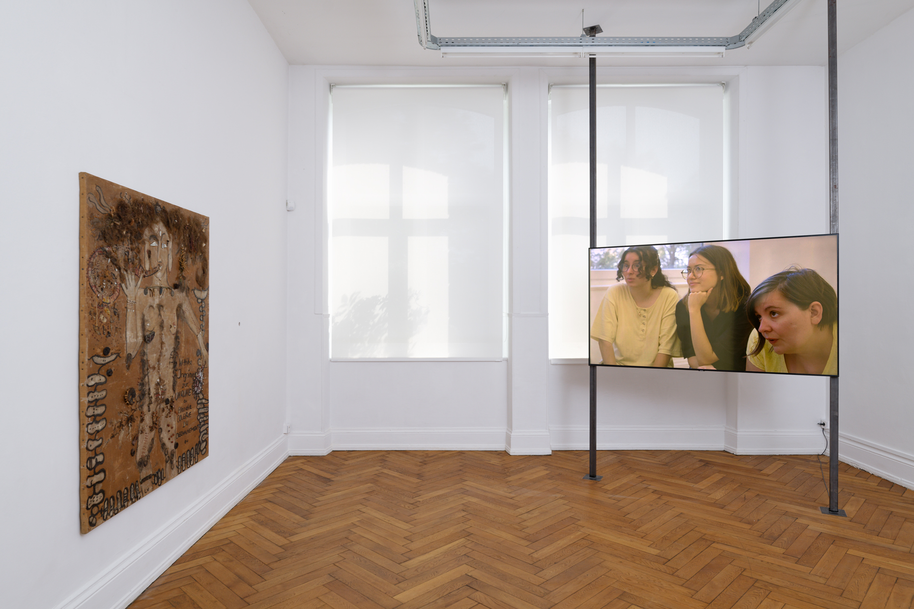 A bright room with wooden floors, white walls, to the left a large collage hangs on the wall and the the left is a screen picturing three young people looking introspectively towards the direction of the collage.