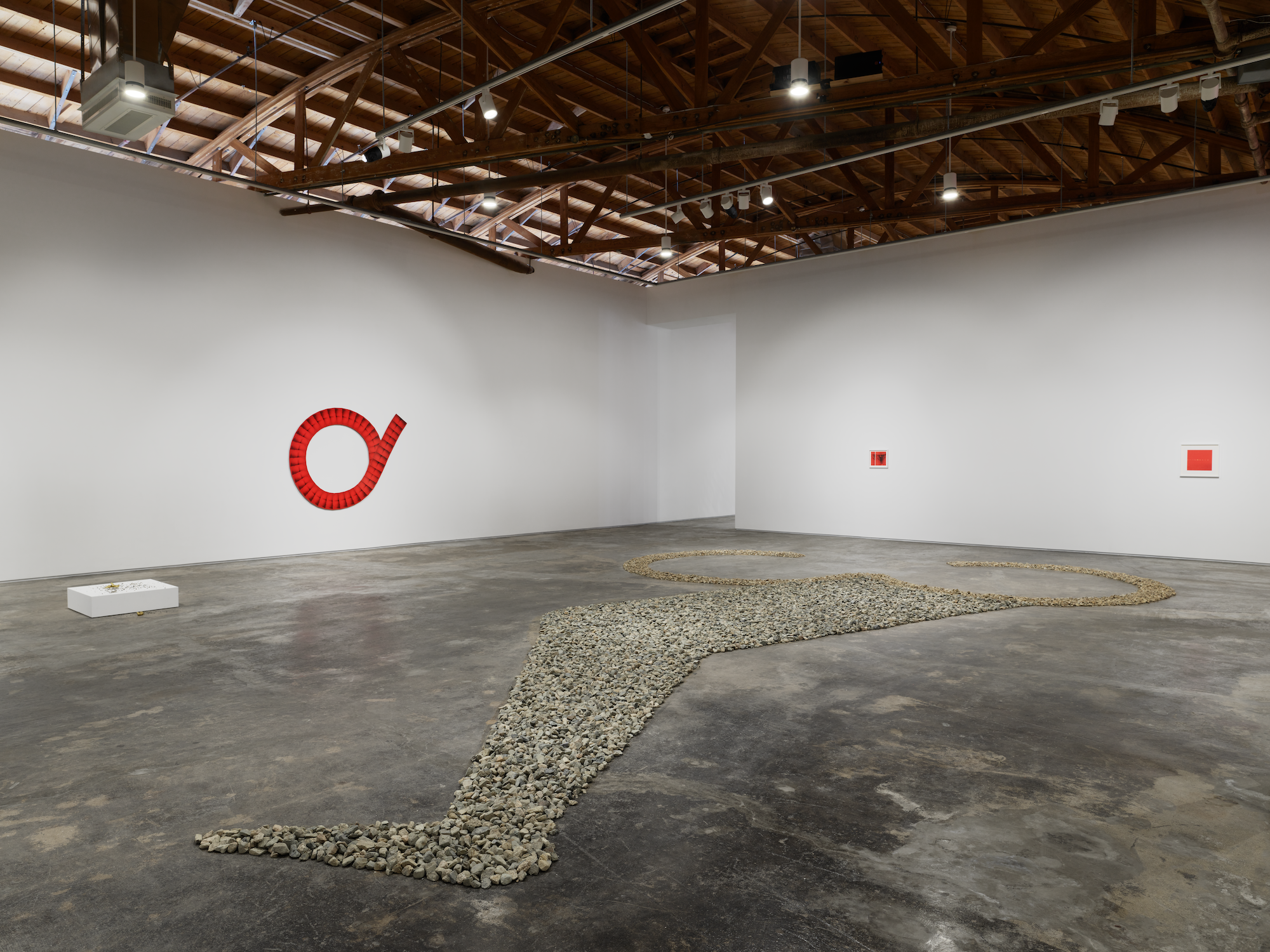 In a large gallery space, hundreds of stones form the shape of a leg on the ground, in the background red prints hang.