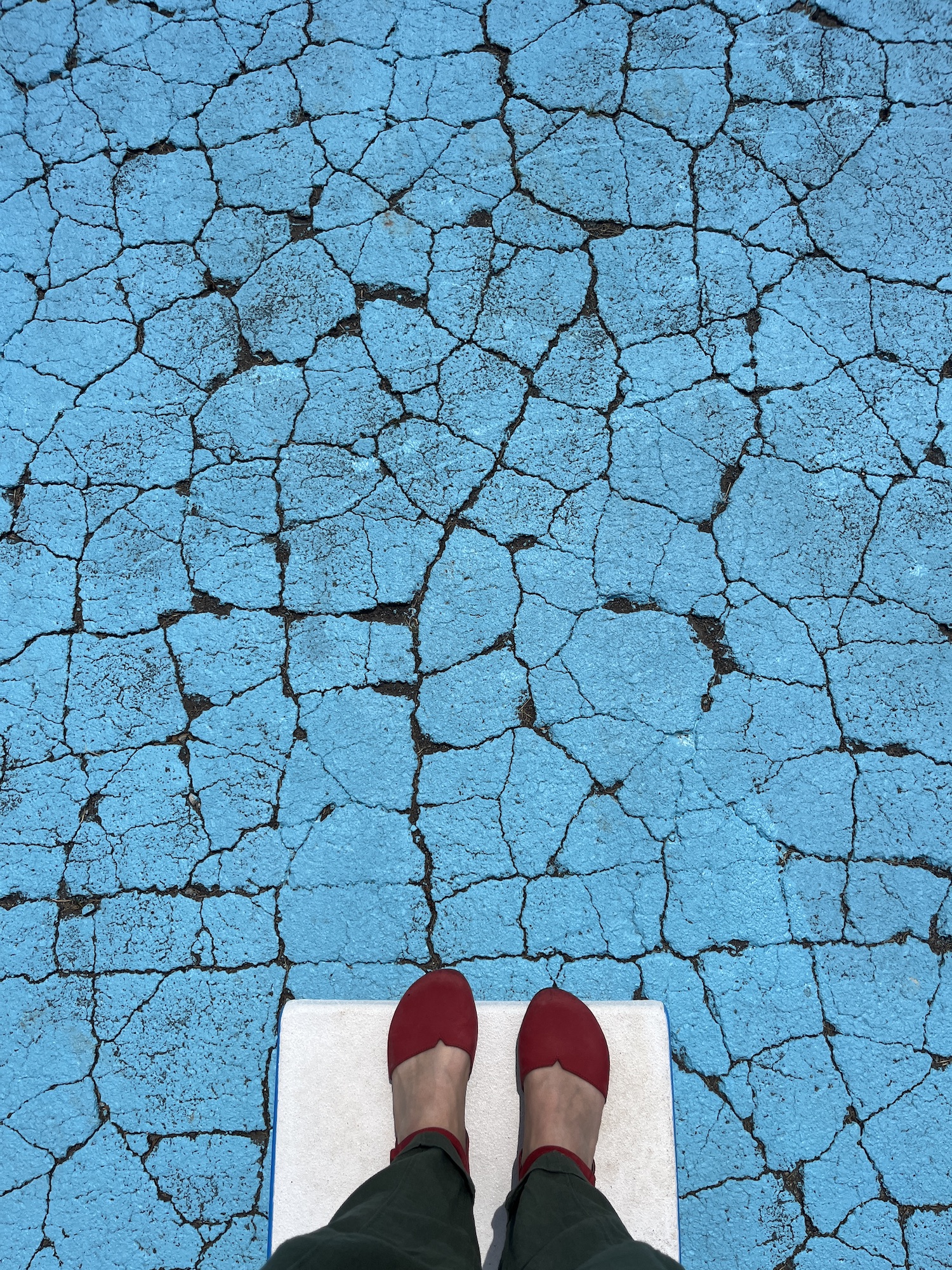 Feet on a diving board lay in the foreground against a light blue background of cracked asphalt.