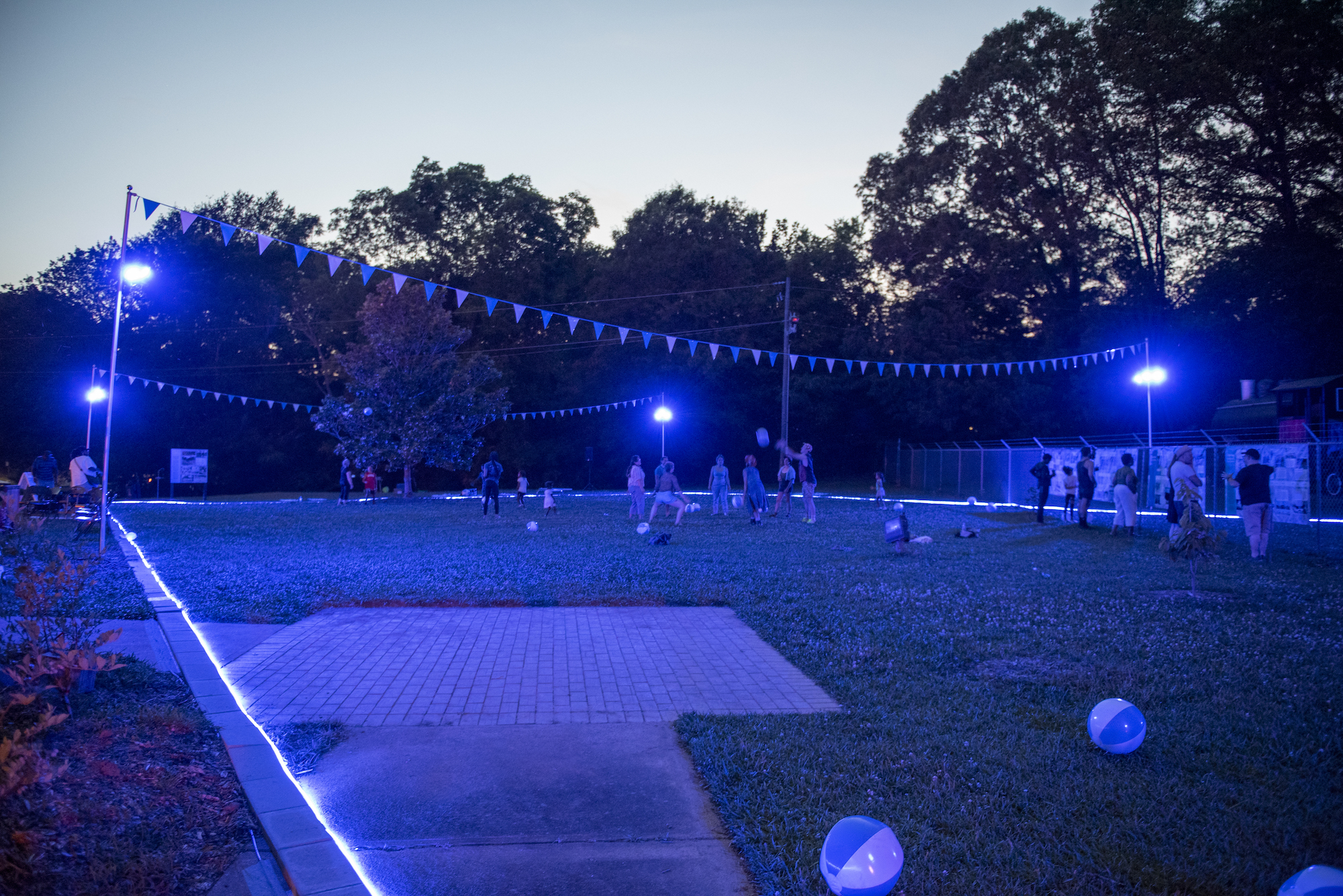 At dusk, many people are gathering in and around a perimeter of a string of blue lights that outline a field where a public pool once was.