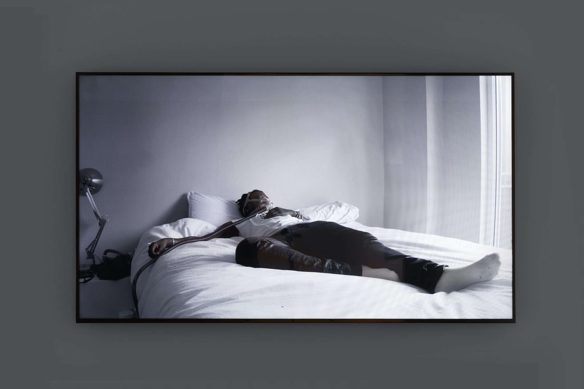 A tv screen displays a video of a person (the artist McClodden) laying on a bed, sprawled out leisurely, with the tendrils of a CPAP machine connected to them.