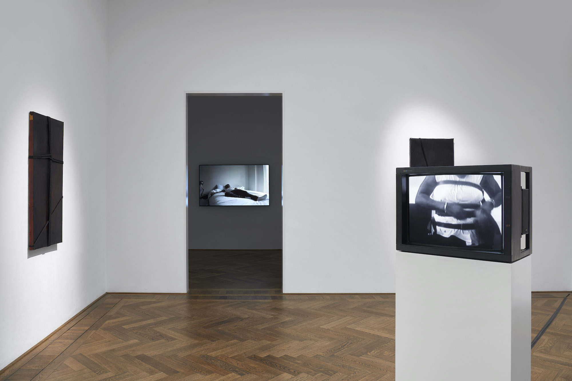 A pristine gallery space with wood herringbone flooring is filled with various sculptures, including two tv monitors displaying videos in black and white.