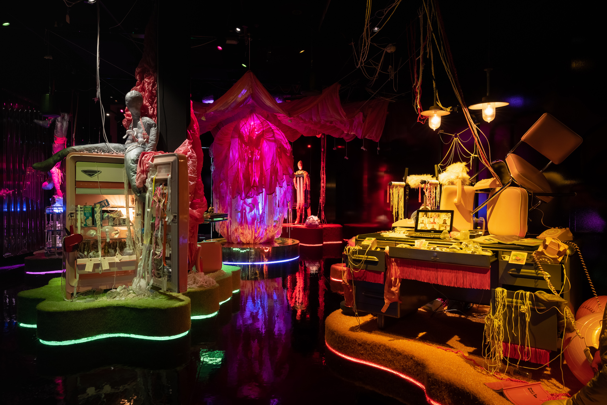 A dark room is lit up with various colorful installations with tassles, fabric and objects spilling out of fridges and a desk; a pink amorphous shape made of fabric hangs in the background.