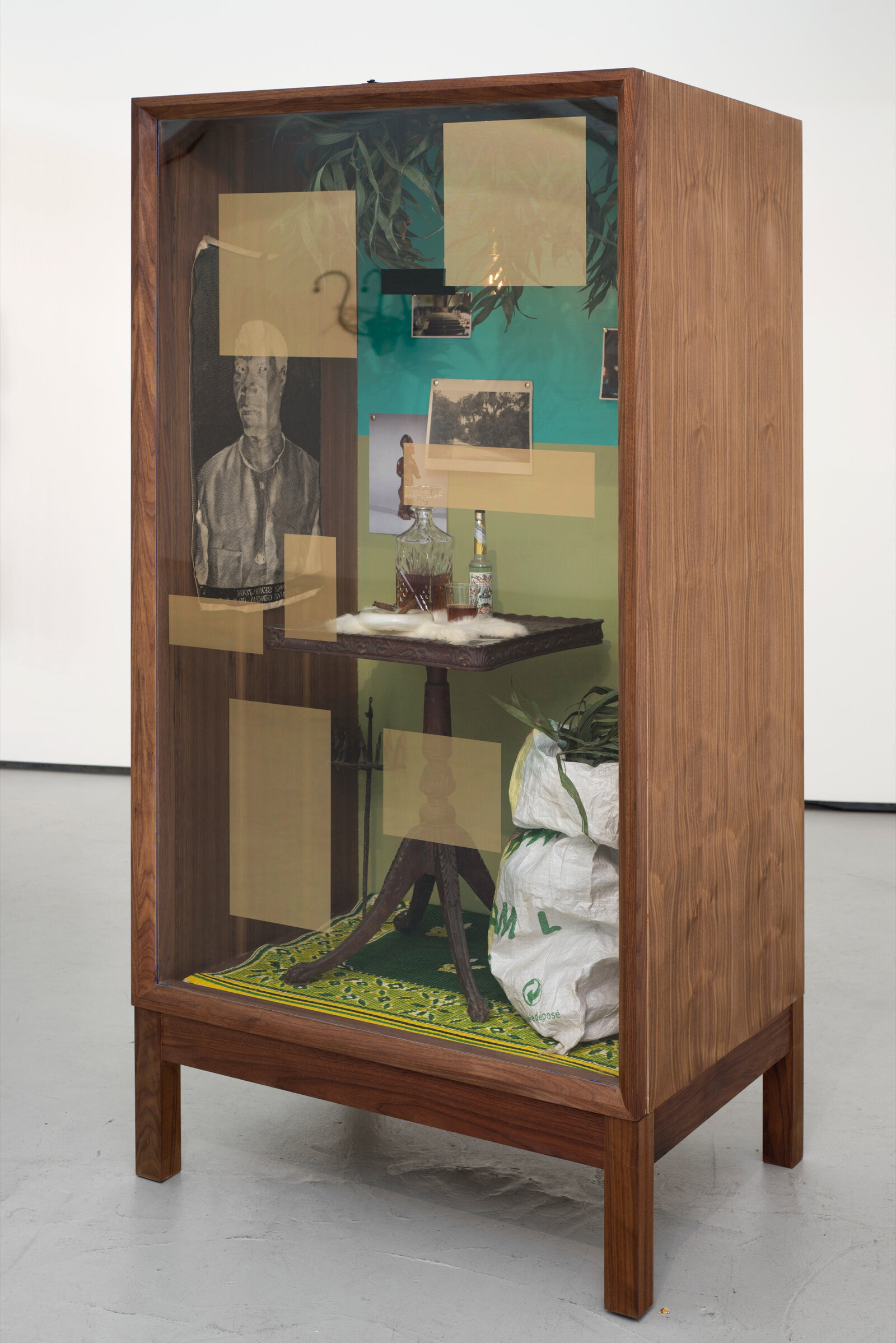 Wood box with glass front on legs. Translucent sawdust colored rectangles on the glass. Behind the glass table a scene with photos tacked to the inside of the box, table with decanter, bottle and glass on top of a small white pelt. Table is on a rug, polypropylene bag of thin tropical looking leaves. Some of the same leaves are hanging from the inside-top of the box as well.