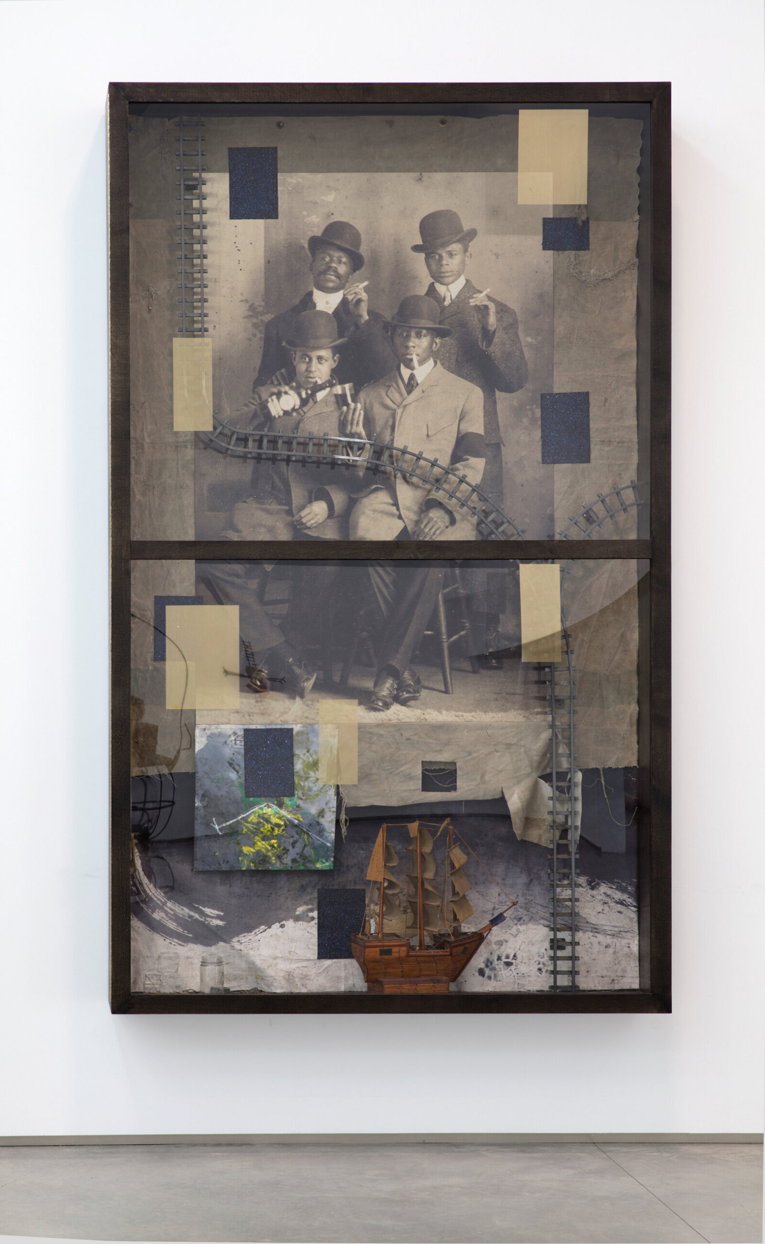 Sepia photo of formally dressed men posing for a portrait smoking cigarettes. Behind photo aged canvas. Tan and Black rectangles, Traint-tracks and model ship assembled around the figures in the photo. One Grey pannel off to the bottom left with yellow and green marks.
