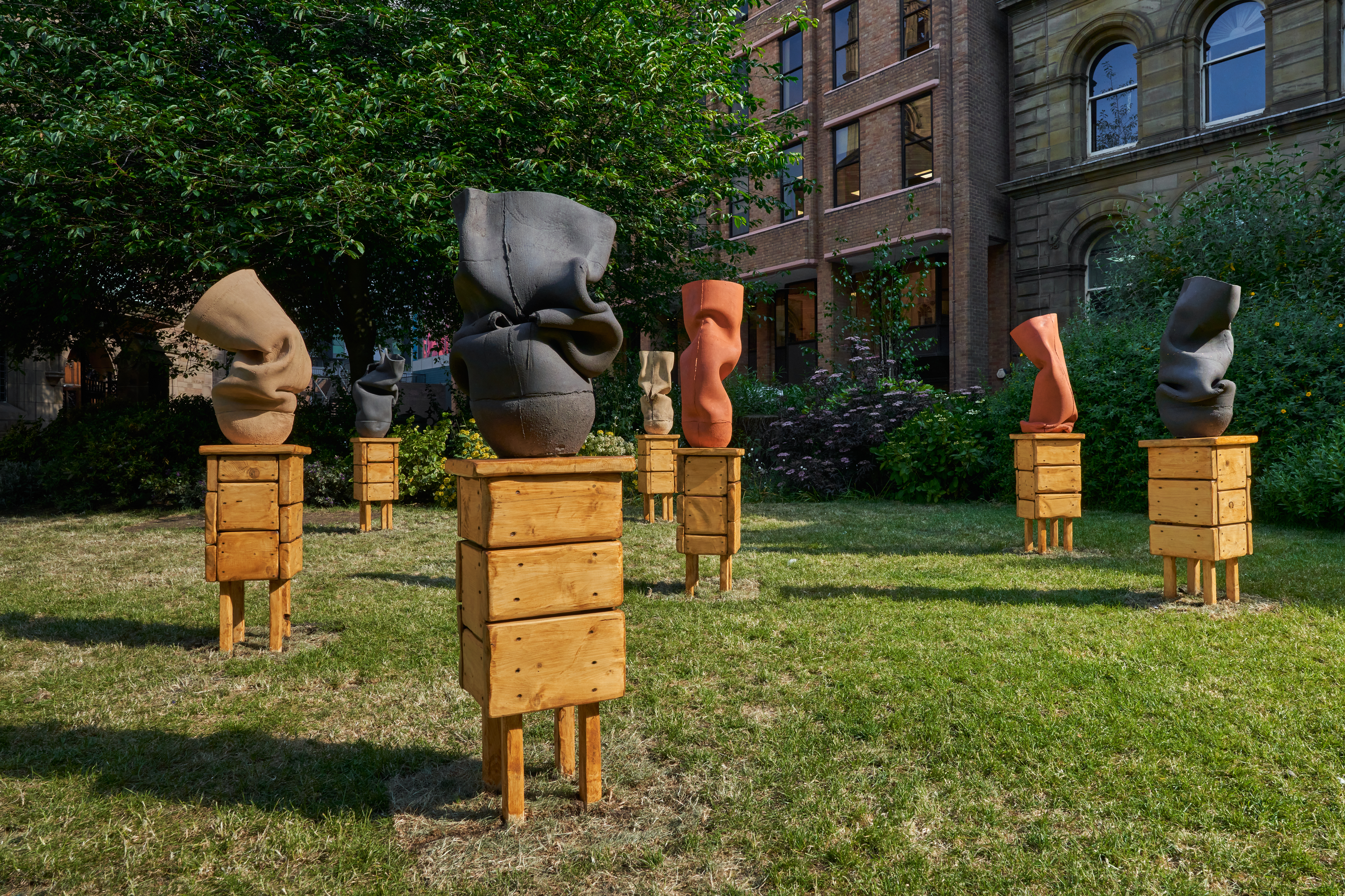 Seven wooden structures holding orange, black, and tan colored sculptural objects are spread out on a green lawn.