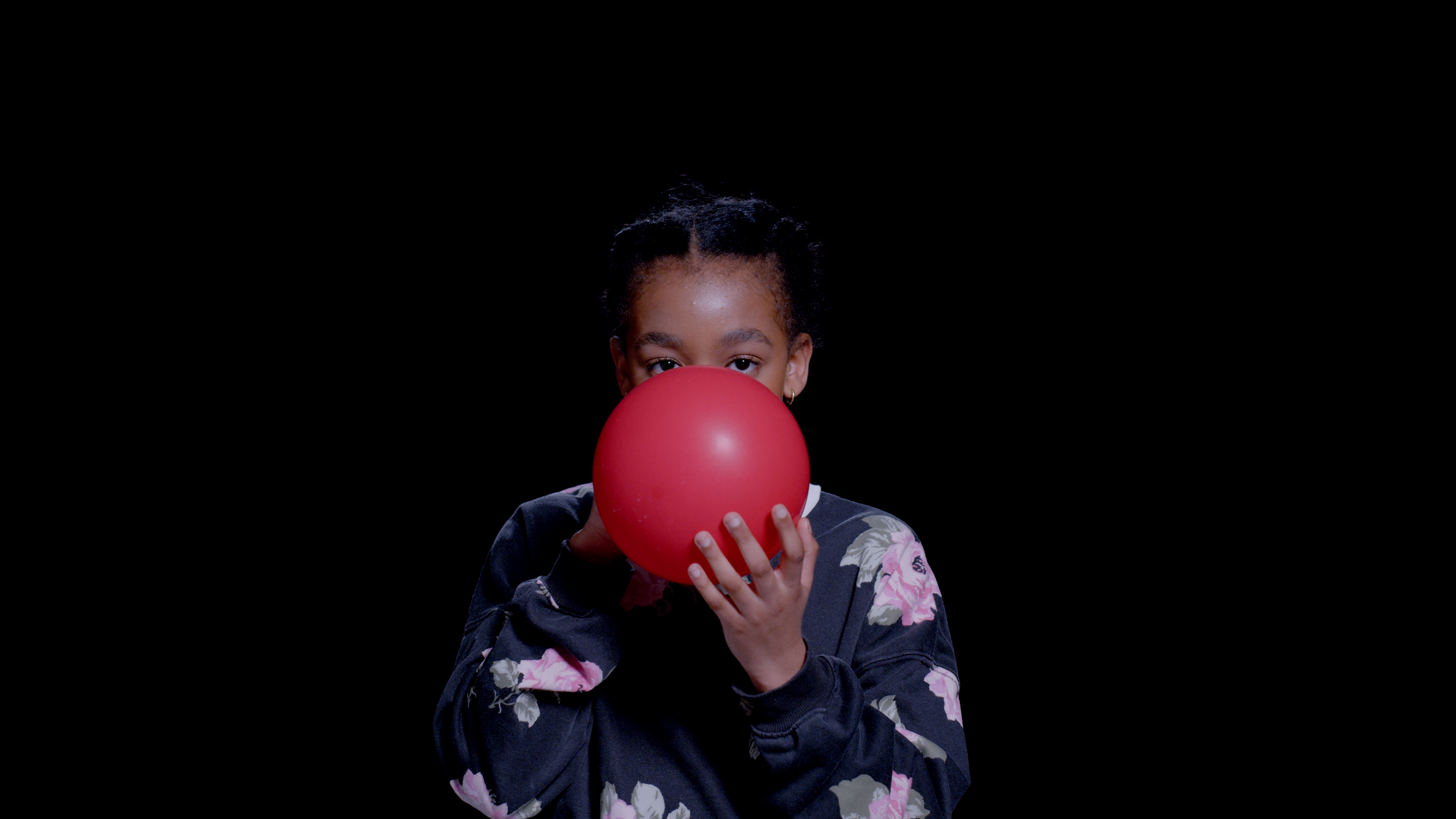 A young black girl in a sweater inflates a red balloon as she stares into the camera.